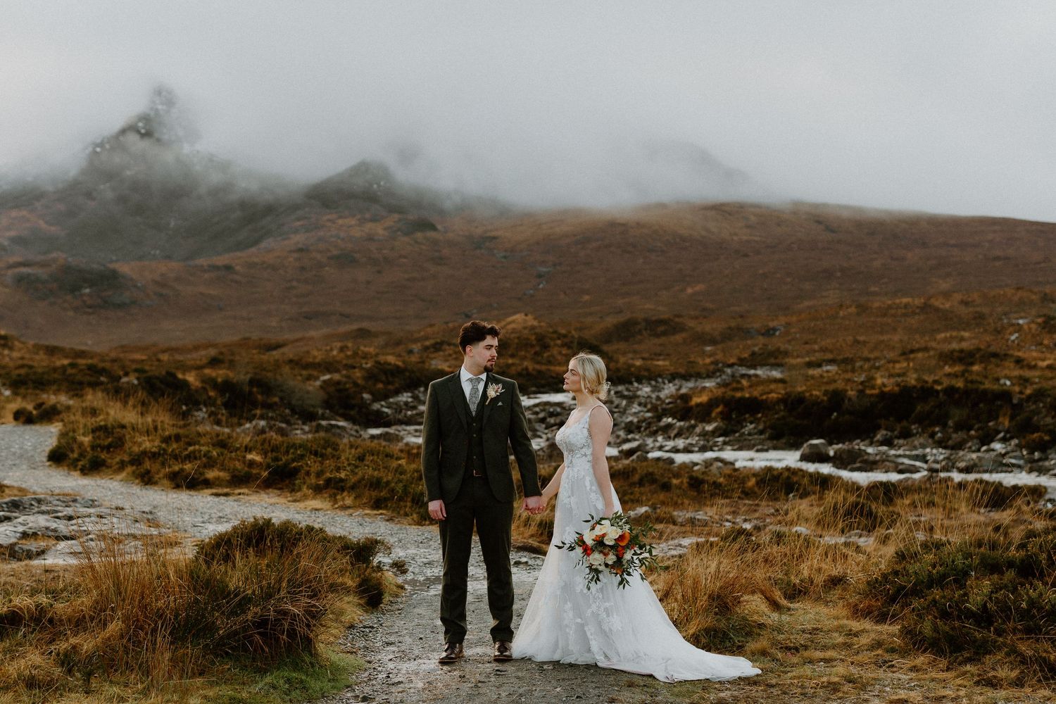 Bride and groom holding hands on a misty mountain path surrounded by dramatic highland scenery.