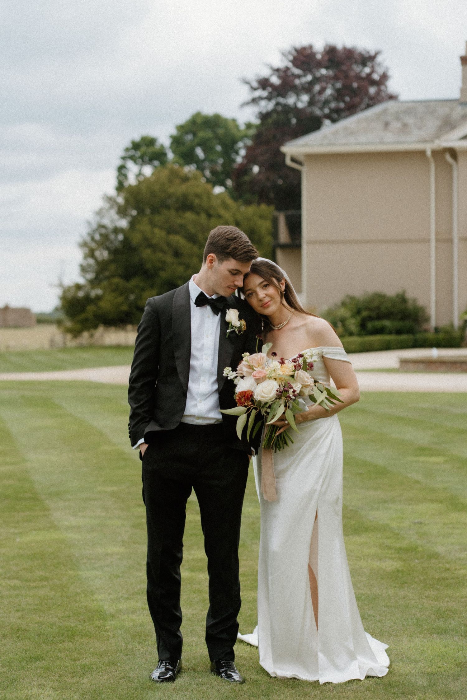 Bride in off-shoulder white gown and groom in black tuxedo sharing intimate moment on manicured lawn.