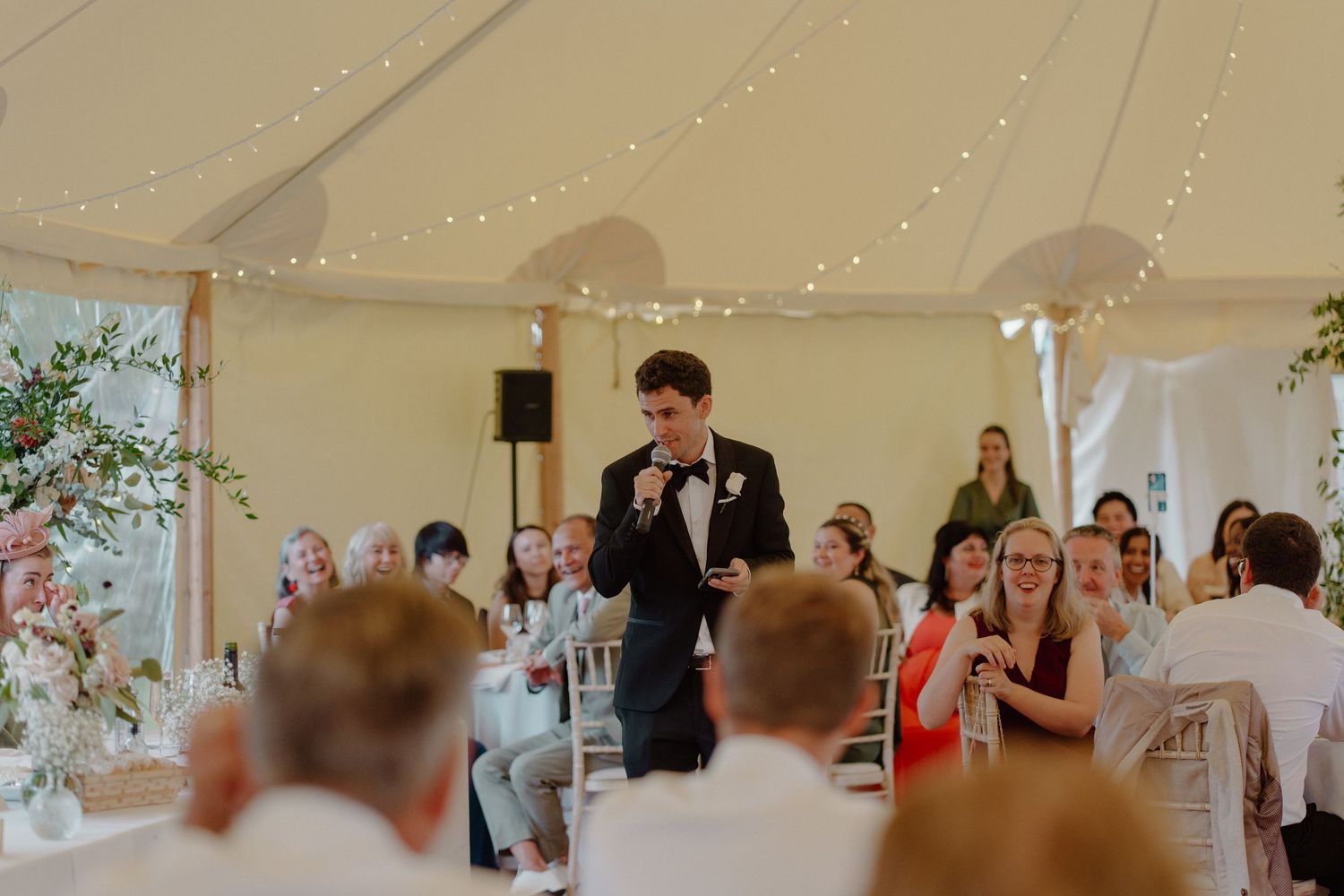 Groom delivering speech at elegant marquee wedding reception with fairy lights.