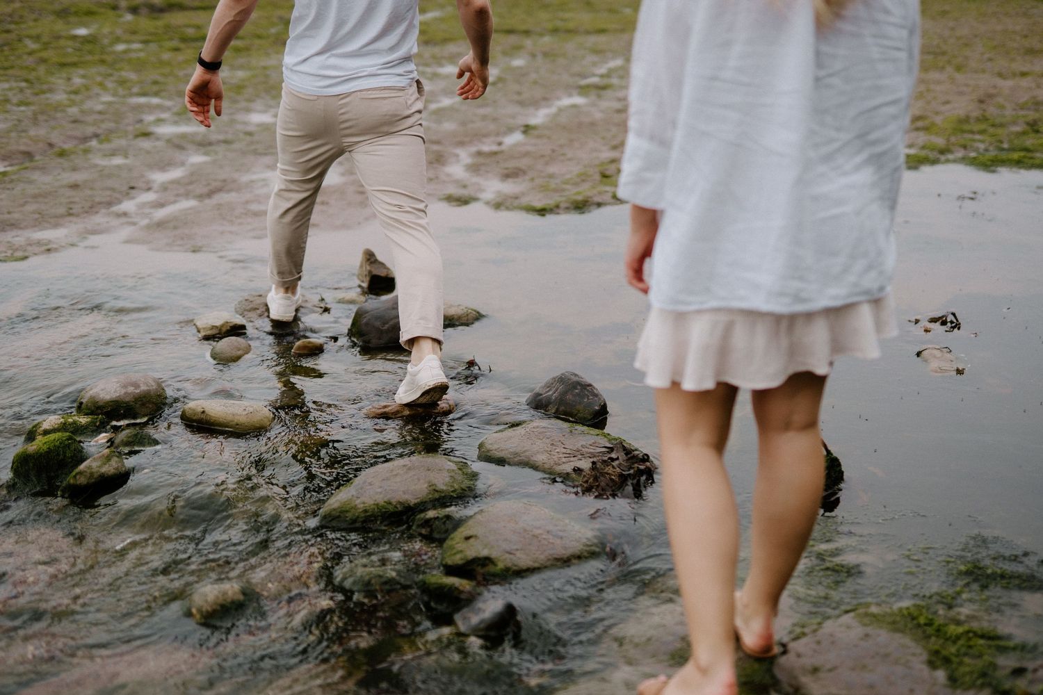 Couple stepping across mossy rocks in shallow water at a scenic coastal location.