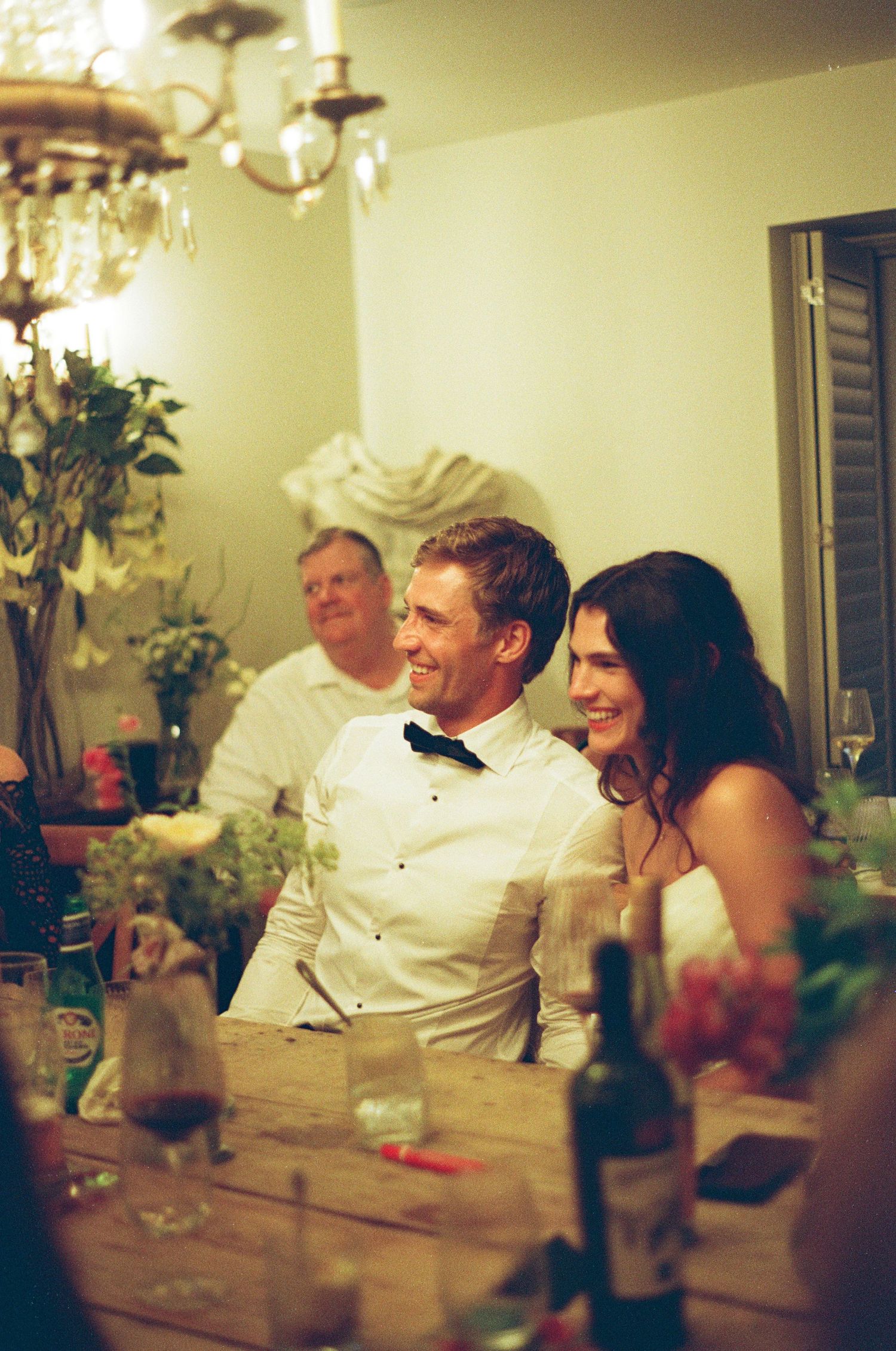 Bride and groom laughing together at wedding reception dinner table beneath elegant chandelier lighting.