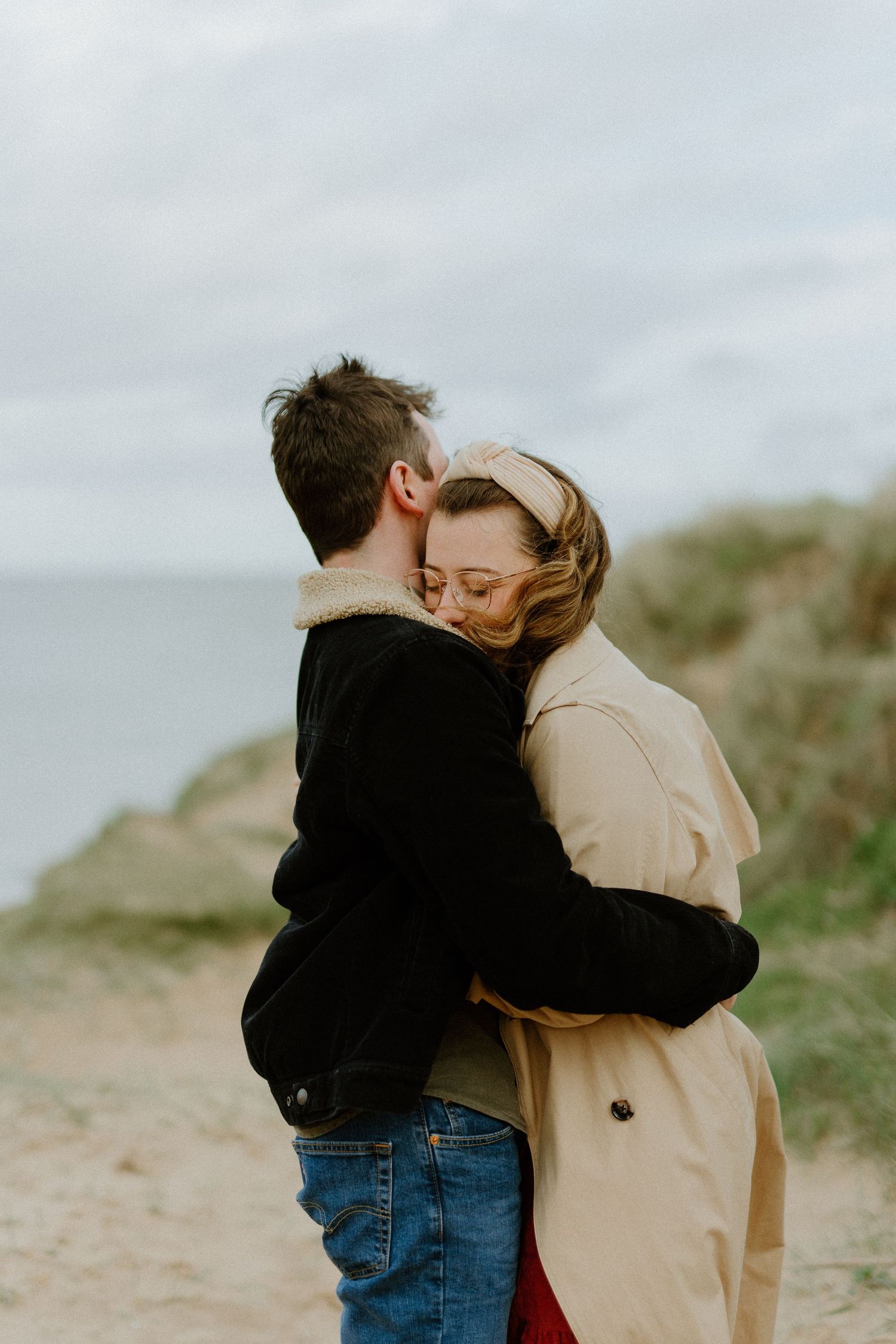 Couple embracing on coastal path, woman in beige trench coat hugging man in black jacket by the sea.