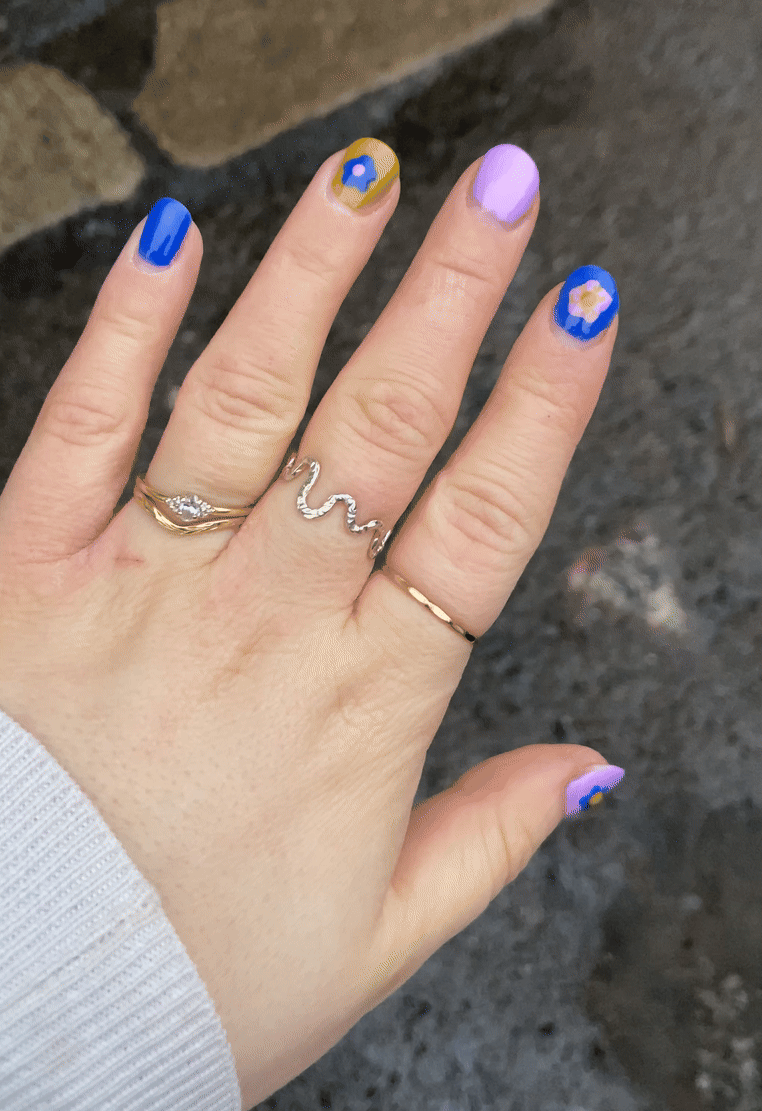 Hand wearing delicate silver rings showcasing bright blue and pink floral nail art design on all fingers.