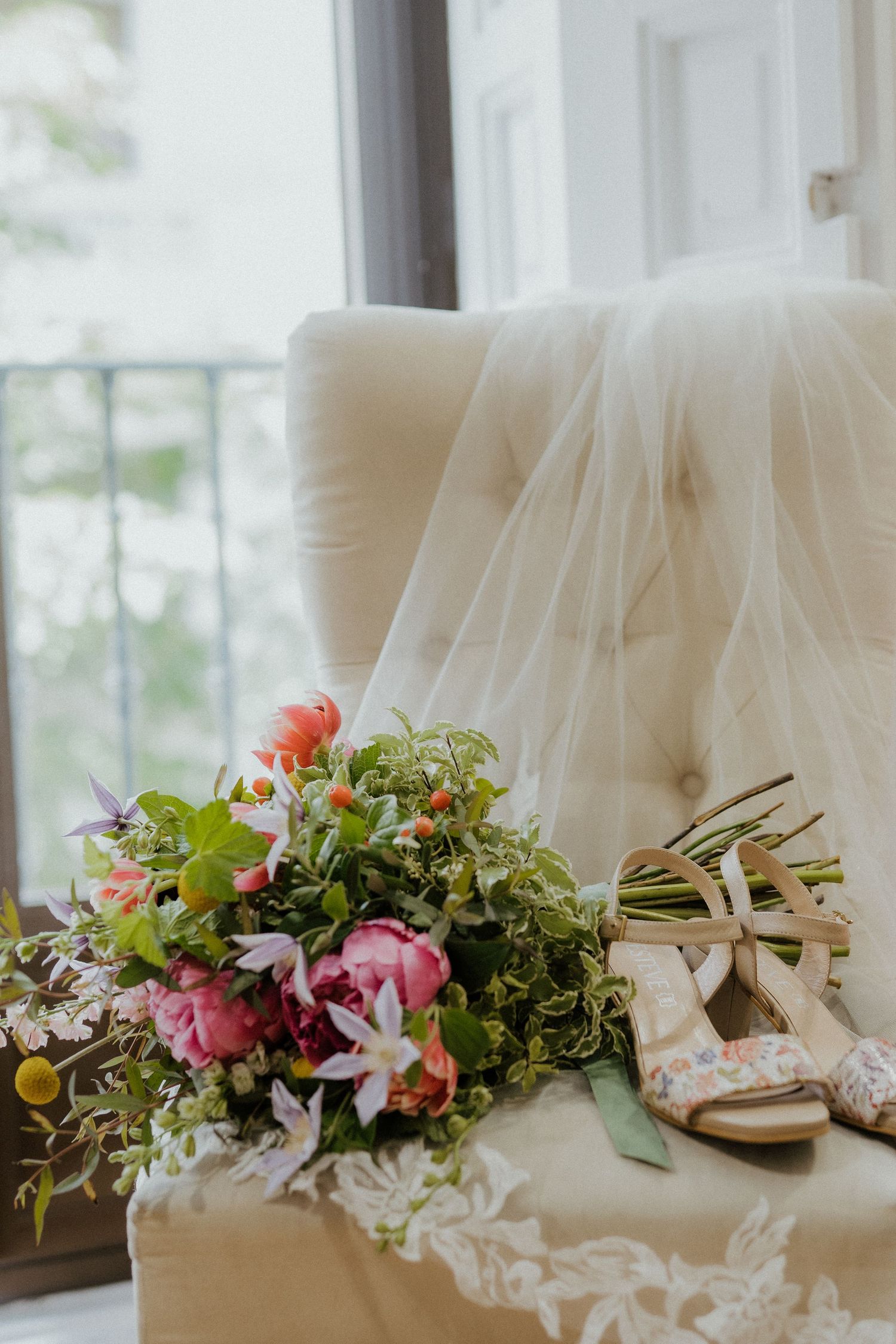 Wedding accessories with bridal veil, floral bouquet and sandals arranged on cream tufted chair by window.