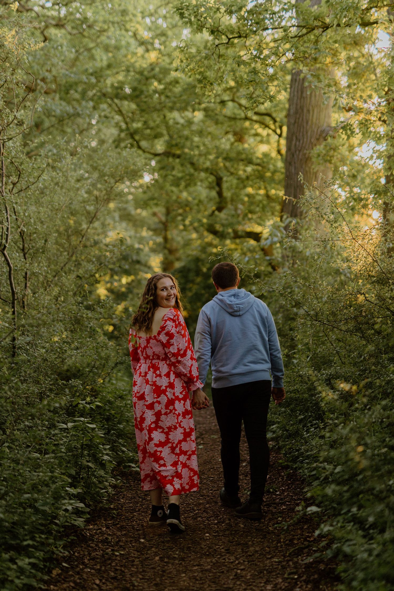 A couple walking together on a forest path, with one wearing a red floral dress and the other in a blue hoodie.