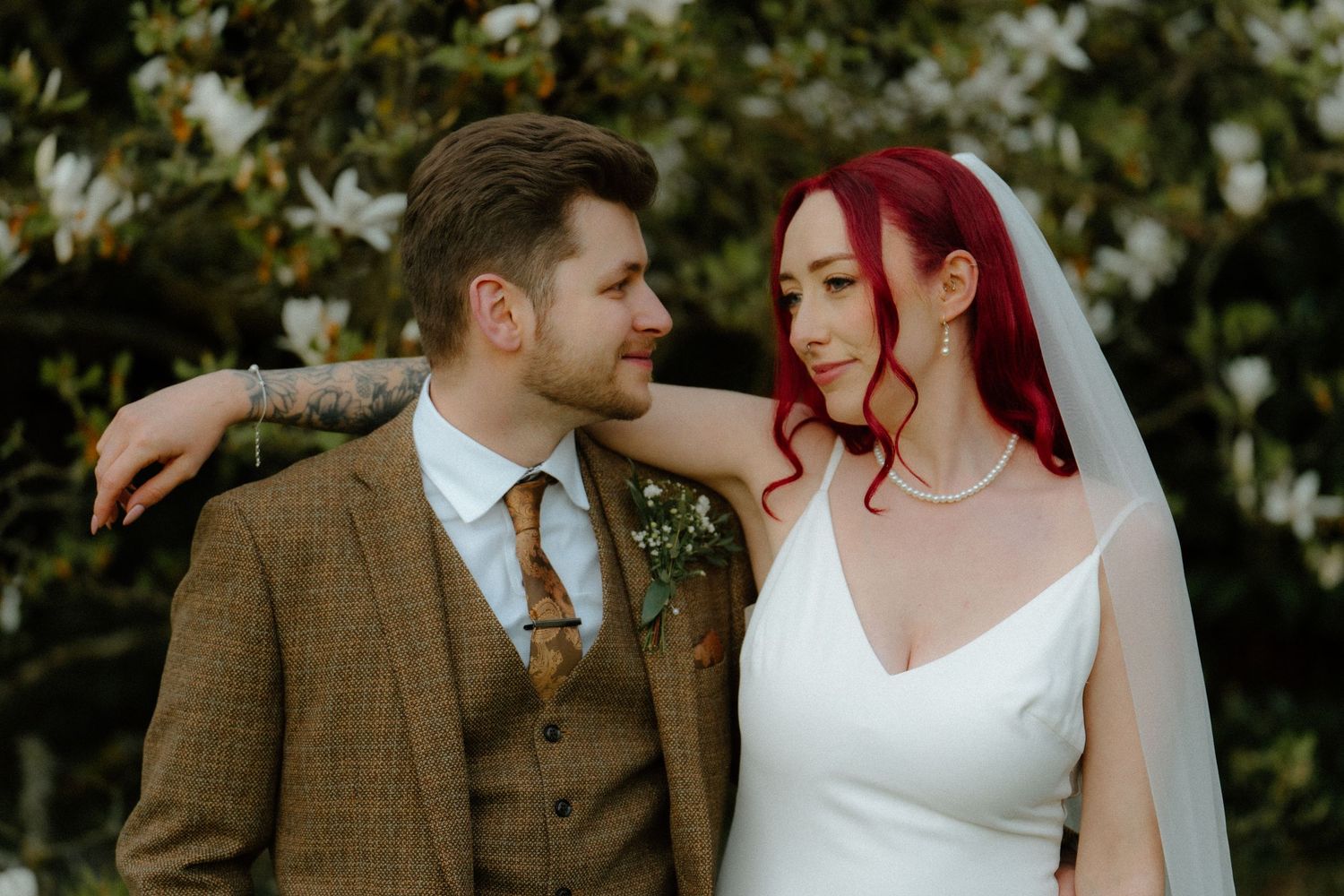 Bride with red hair and groom in brown tweed suit pose together outdoors near white flowers on their wedding day.