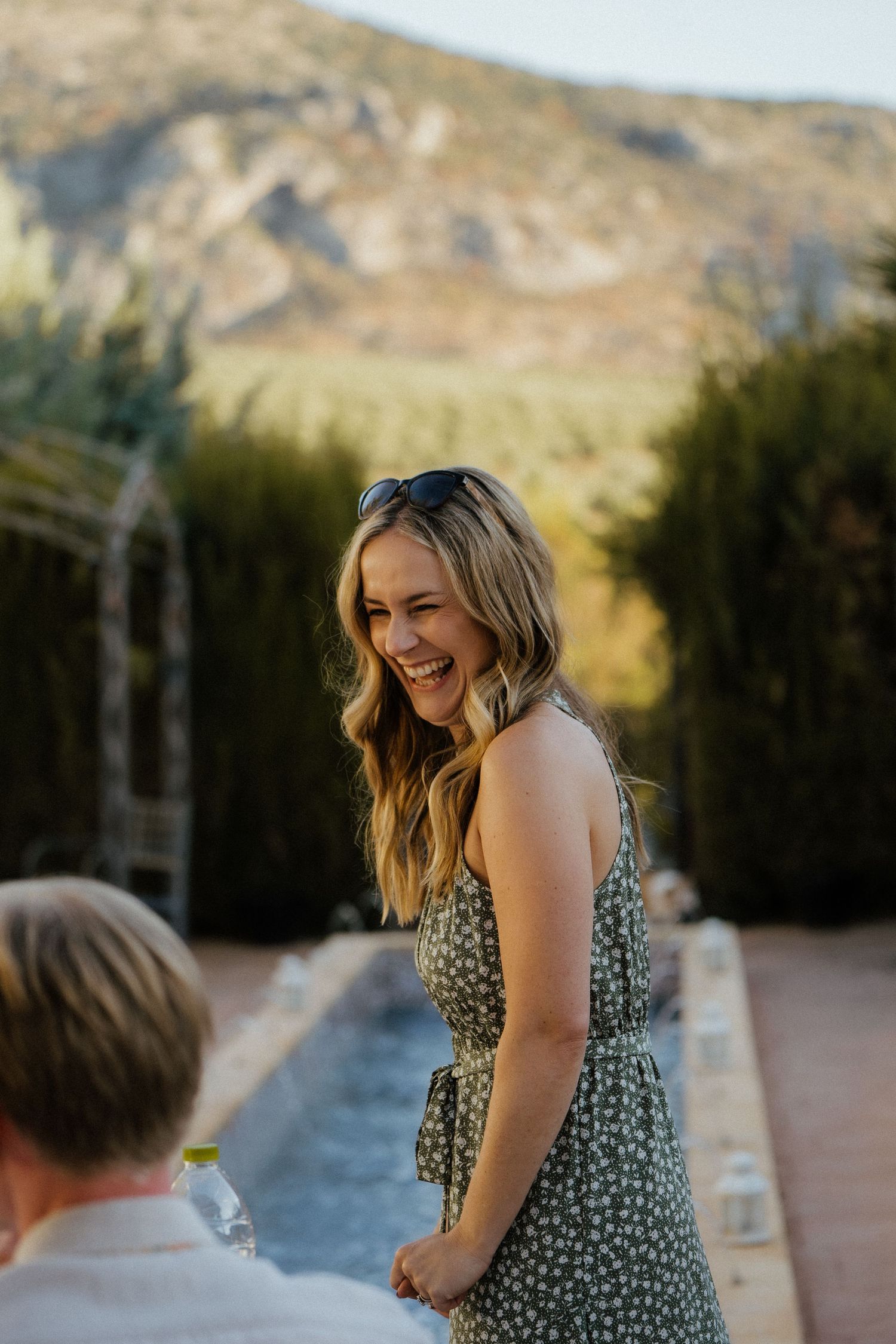 Joyful woman in green floral sundress laughing at outdoor wedding reception with mountain backdrop.