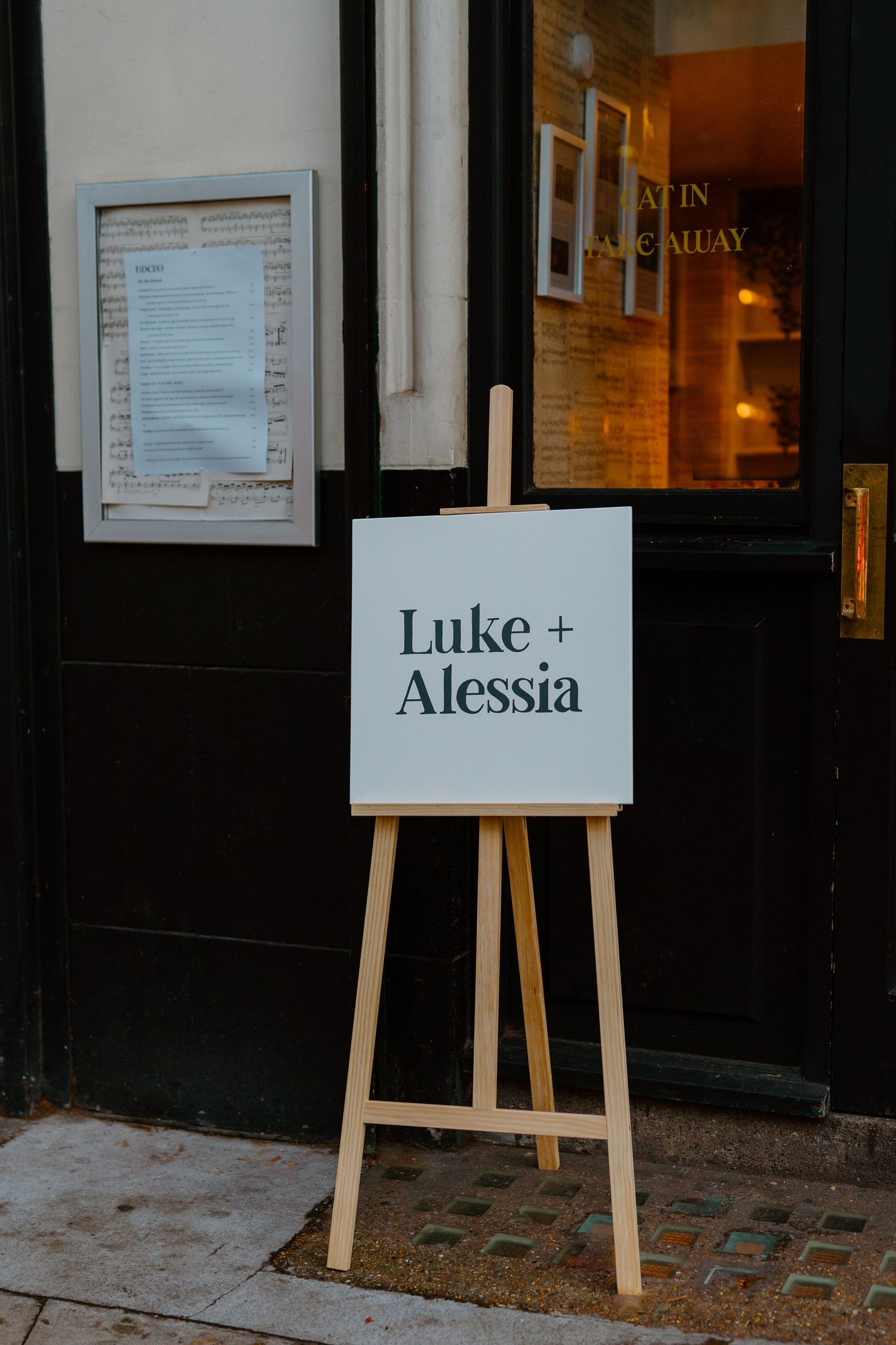 Wooden easel displaying a white wedding welcome sign reading Luke and Alessia outside a venue entrance.