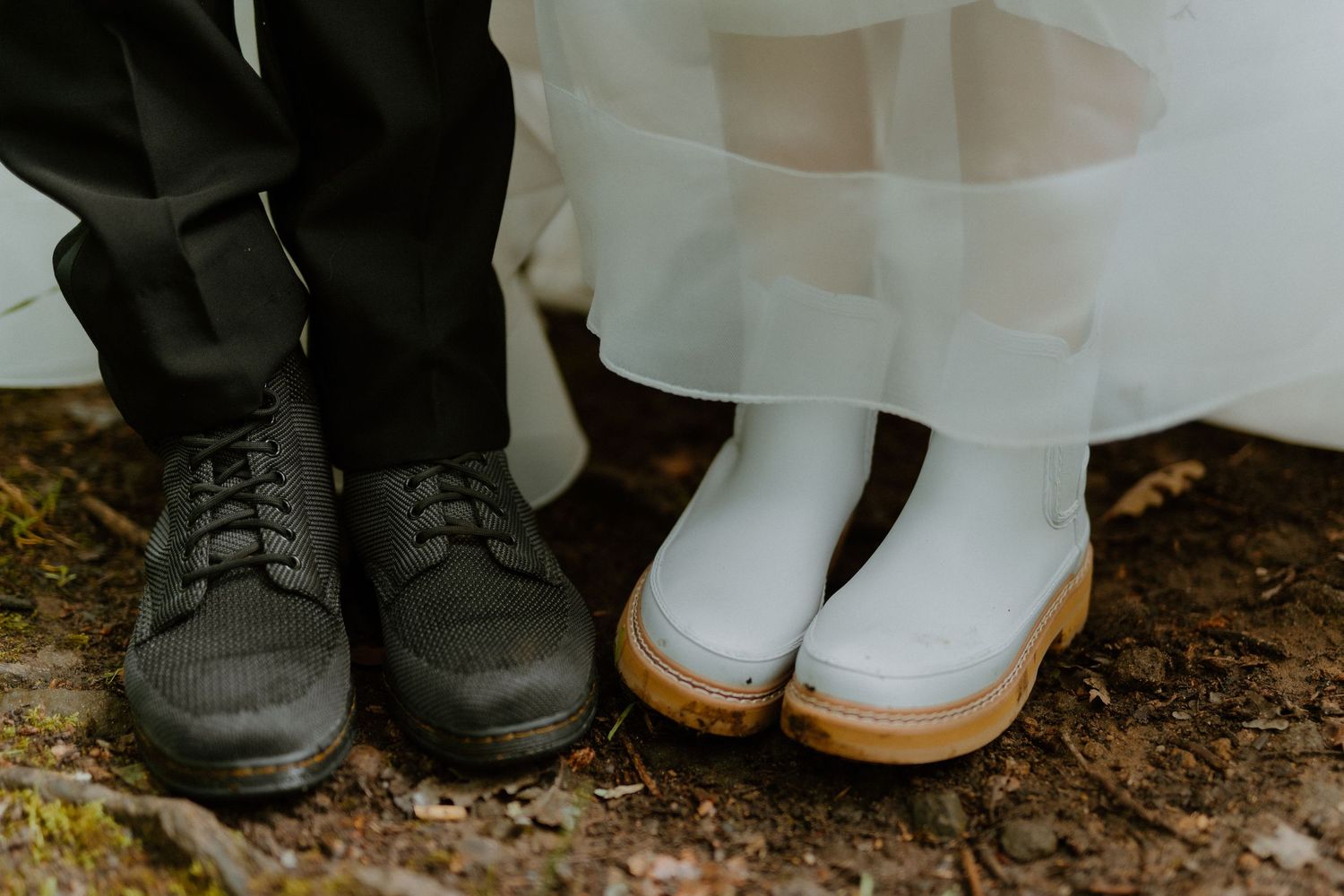 Close-up of bride in white rubber boots and groom in black dress shoes standing outdoors.