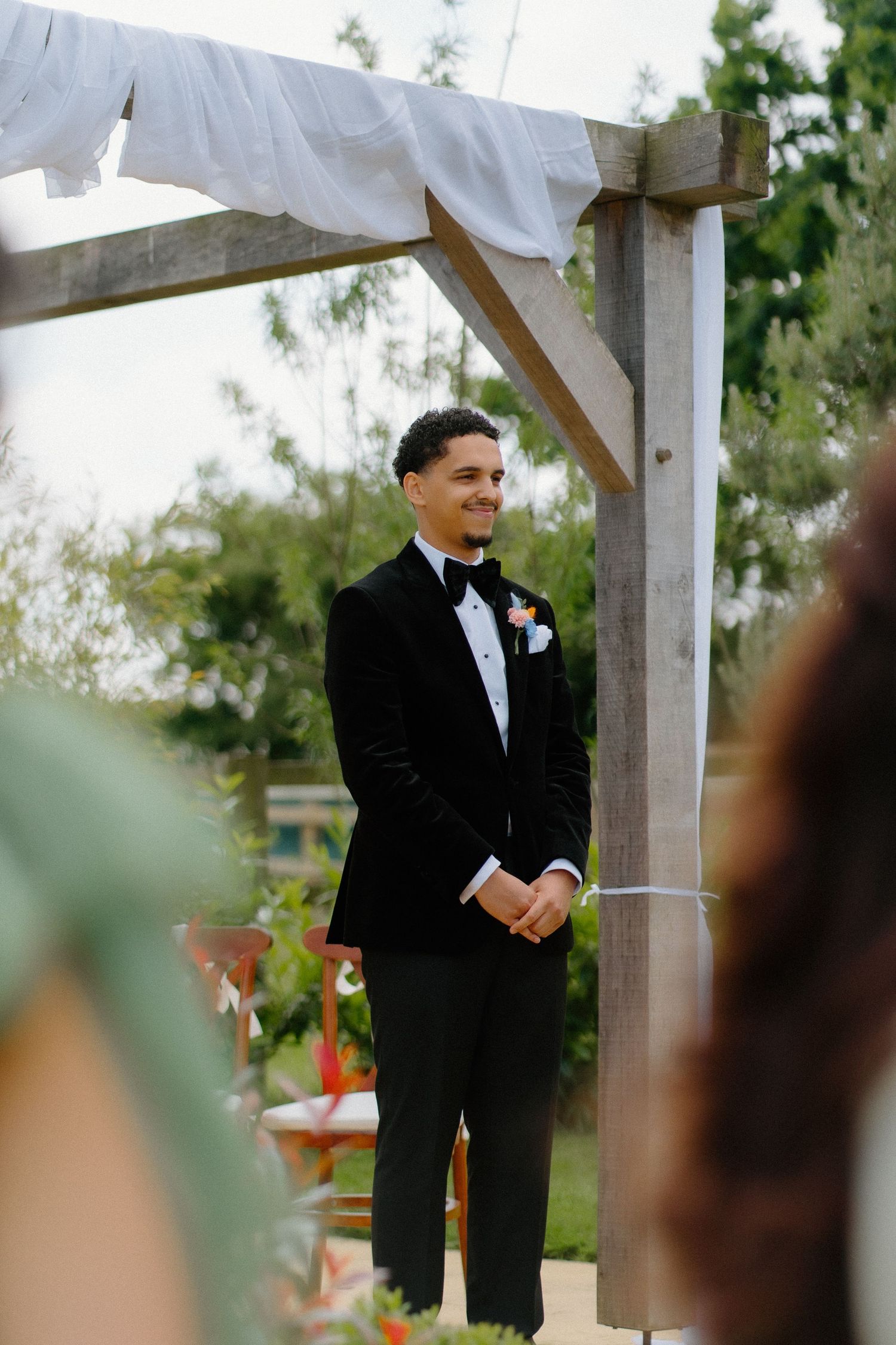 Groom in black tuxedo smiling warmly under a wooden arch as bride approaches during outdoor ceremony.