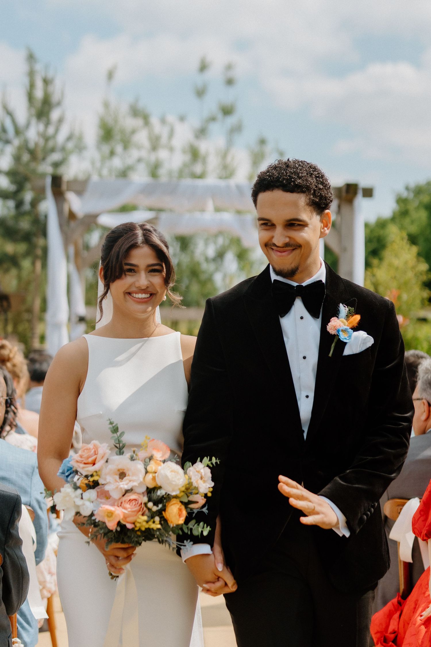 Newlywed couple walks down aisle smiling, bride holding pastel bouquet and groom in black velvet tuxedo.
