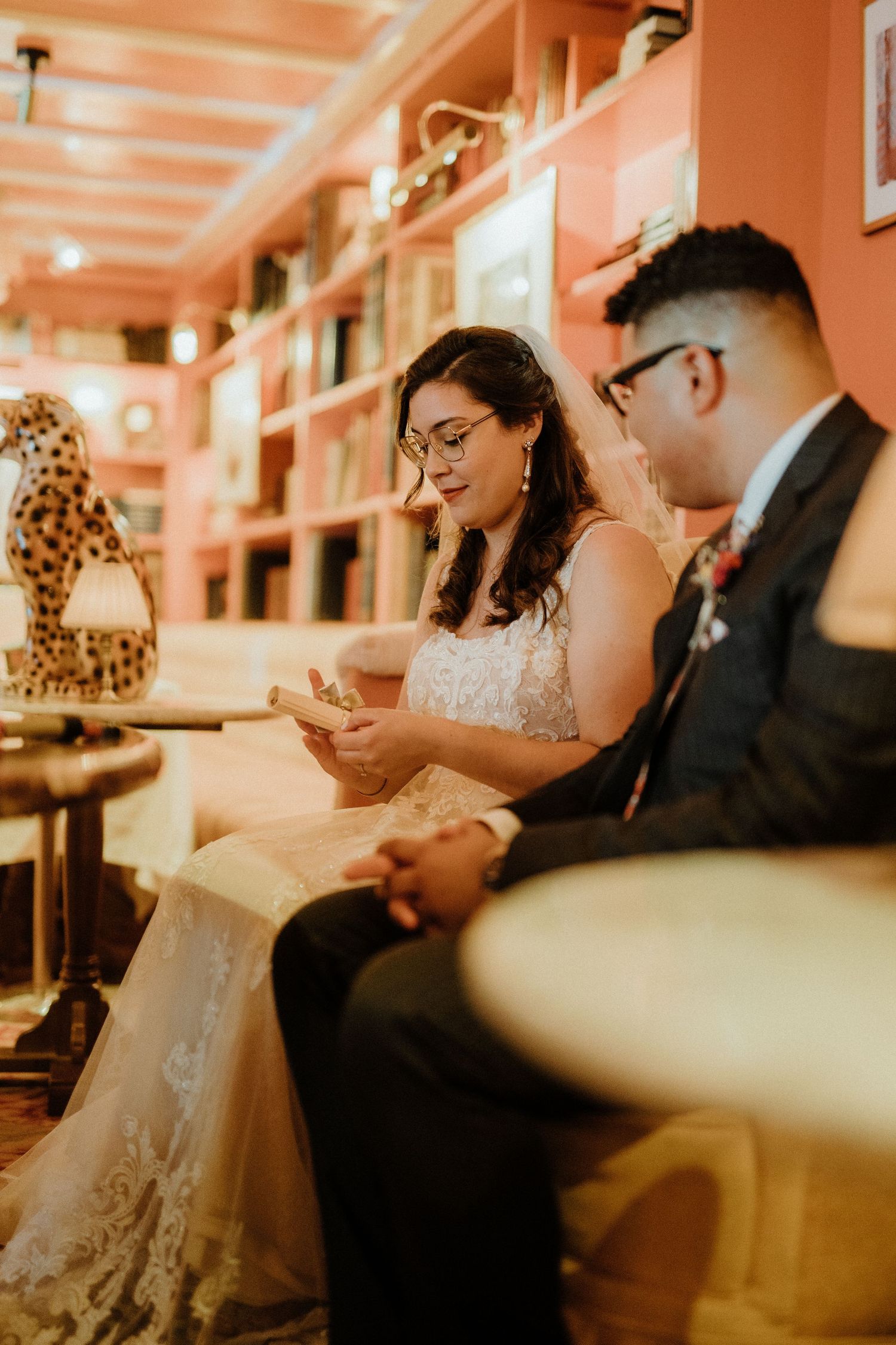 Bride and groom in glasses sit together on sofa reading vows in warm pink room.