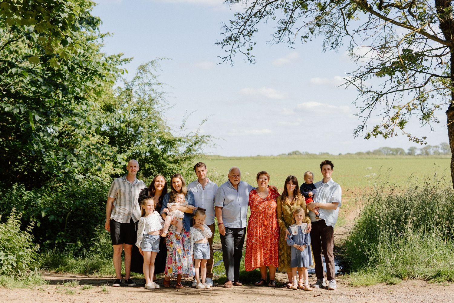 Large multigenerational family posing together on a sunny countryside path with green fields.