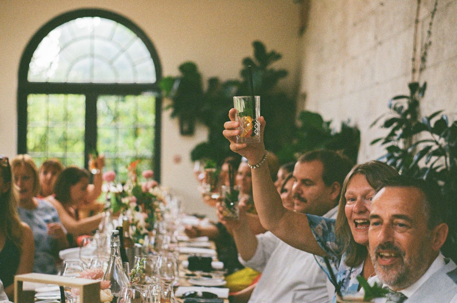 Wedding guests raise champagne glasses in joyful toast at elegant reception dinner table with lush greenery backdrop.
