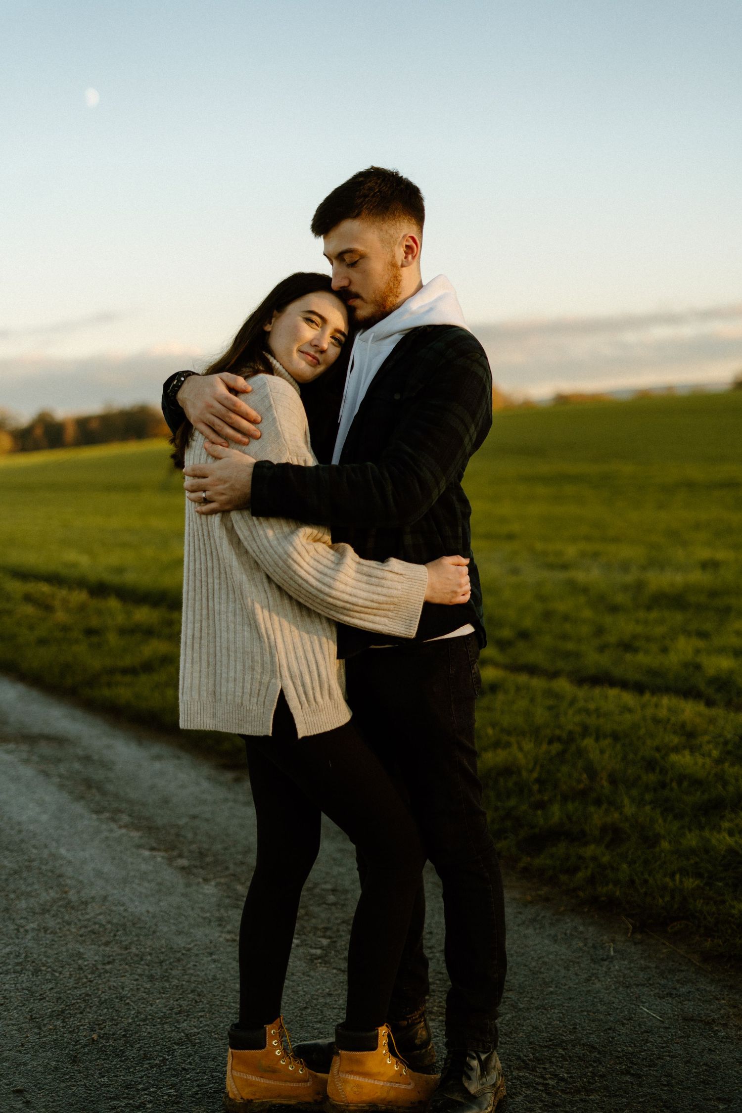 Couple embracing on countryside path at golden sunset with green fields in background.