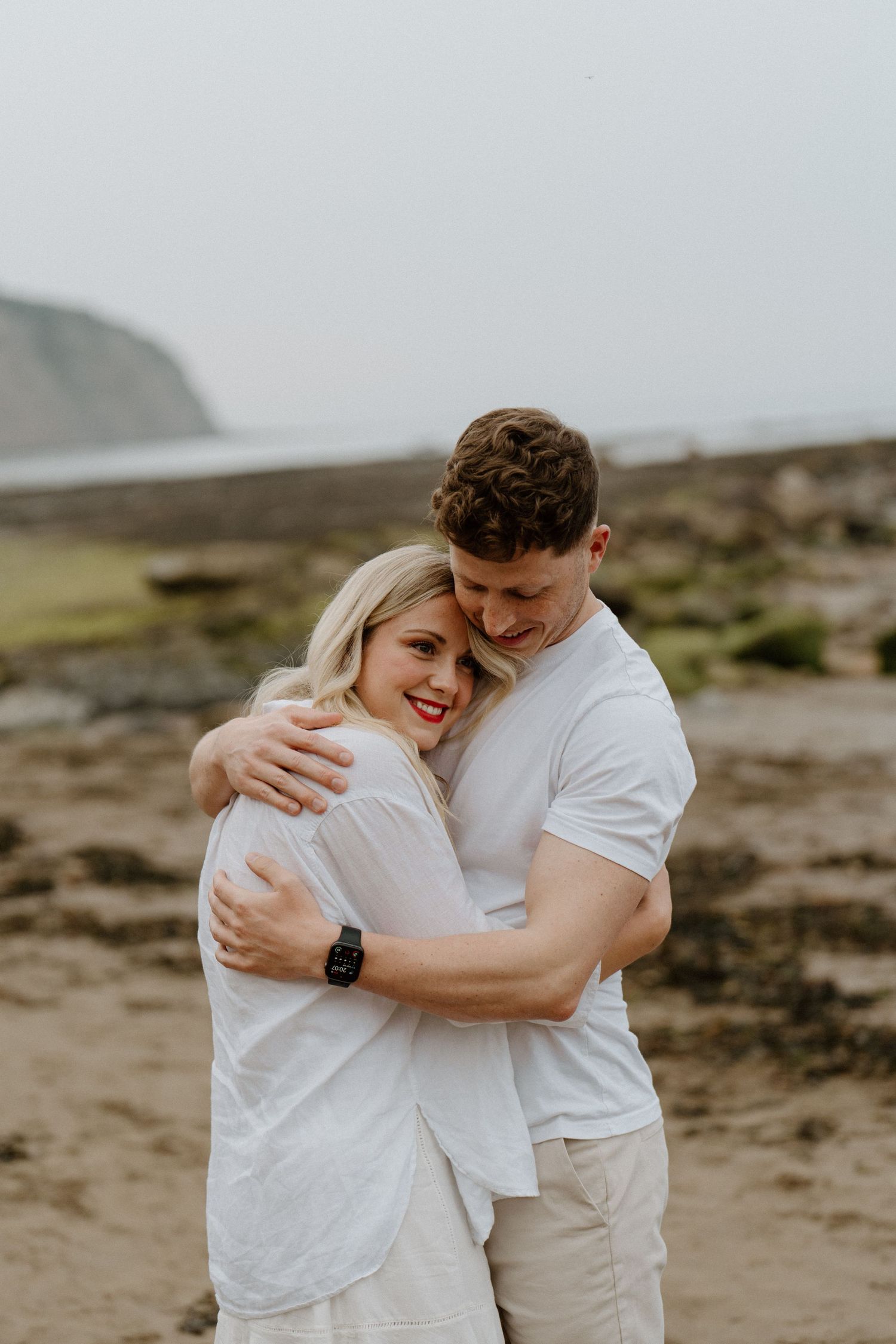Couple sharing a warm embrace on a rocky beach with coastal cliffs visible in the background.