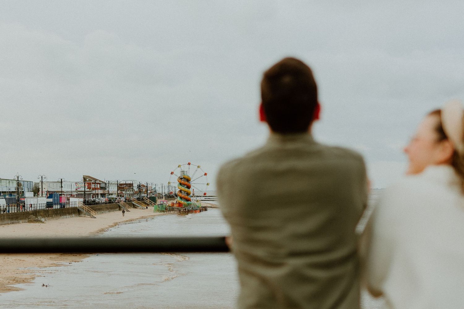 Couple leaning on railing overlooking a seaside town with a Ferris wheel in background.