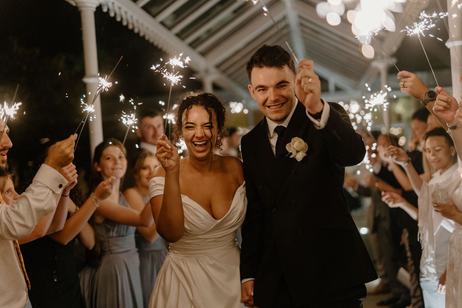 Smiling bride and groom walk through a sparkler send-off with joyful wedding guests.