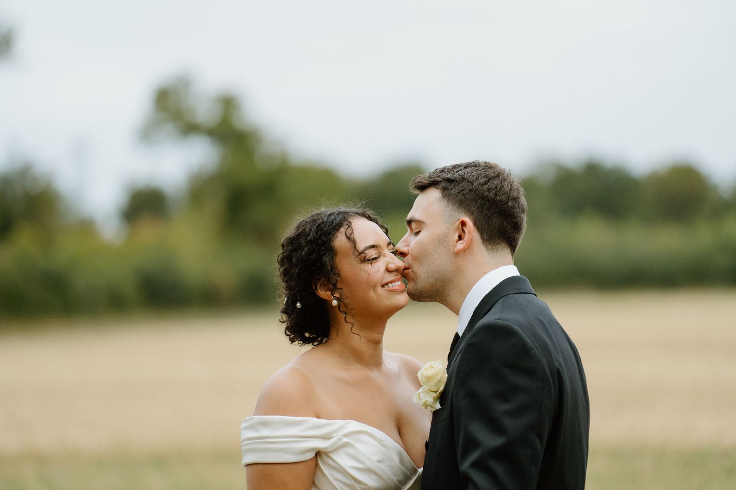 Groom kisses bride on cheek in romantic outdoor wedding portrait in golden field setting.