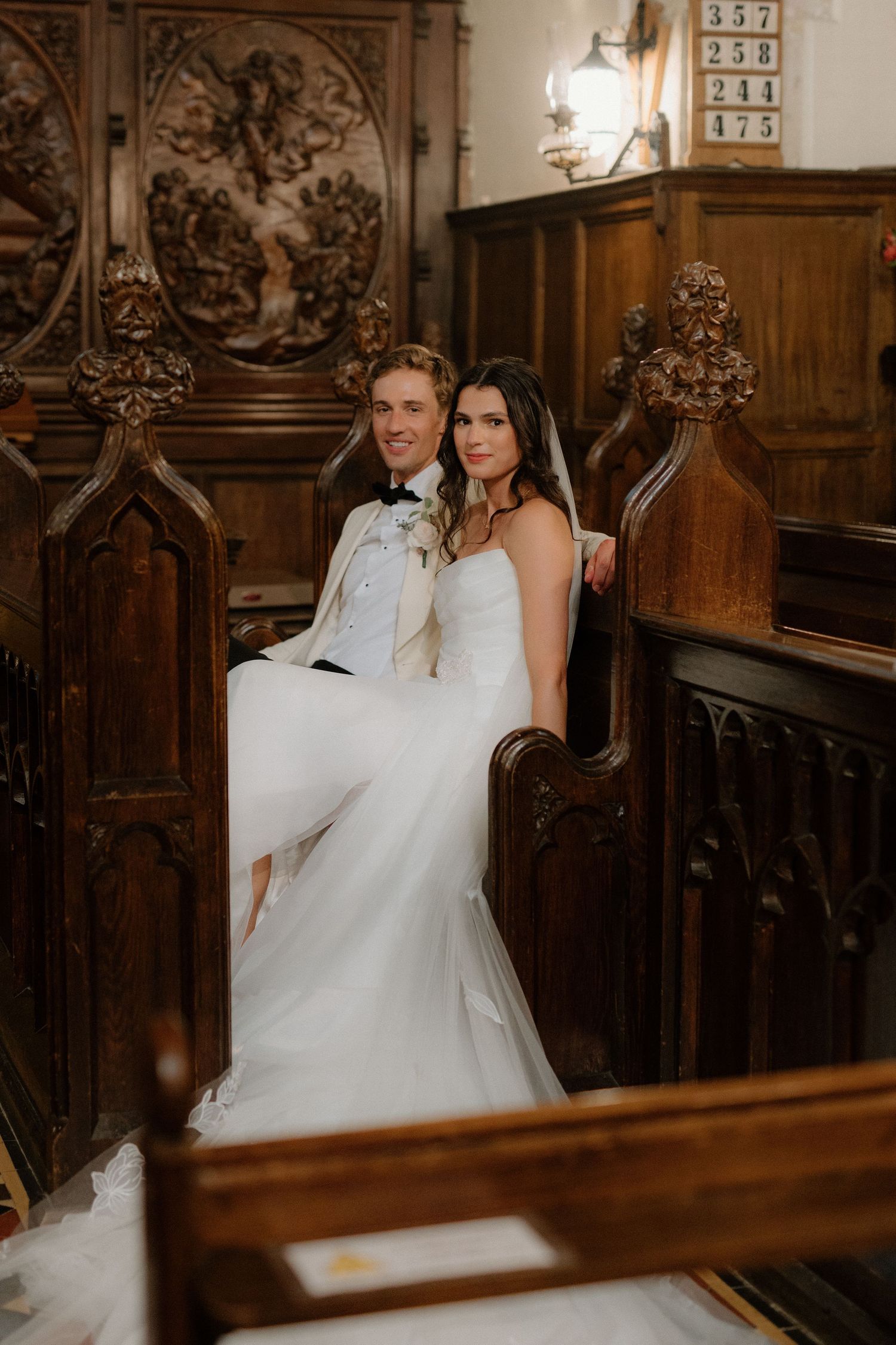 Bride and groom sit together in ornate wooden church pews, smiling warmly in a richly decorated interior.