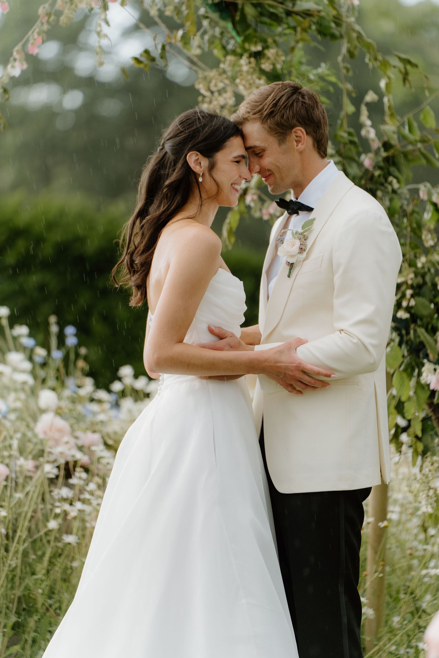Smiling bride and groom embracing under floral arch surrounded by pink and white wildflowers.