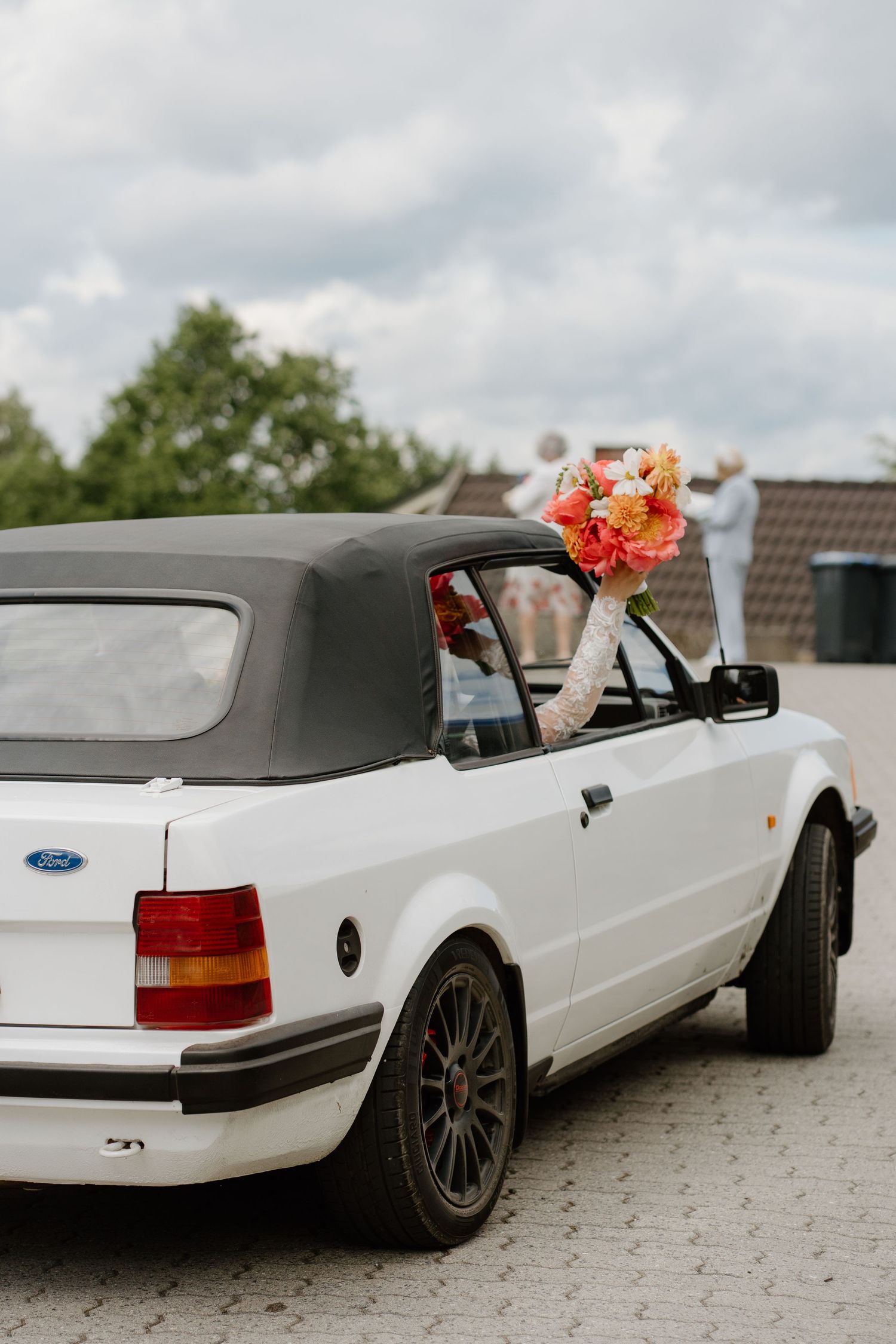 Bride holding colorful bouquet out window of white Ford convertible after wedding ceremony.