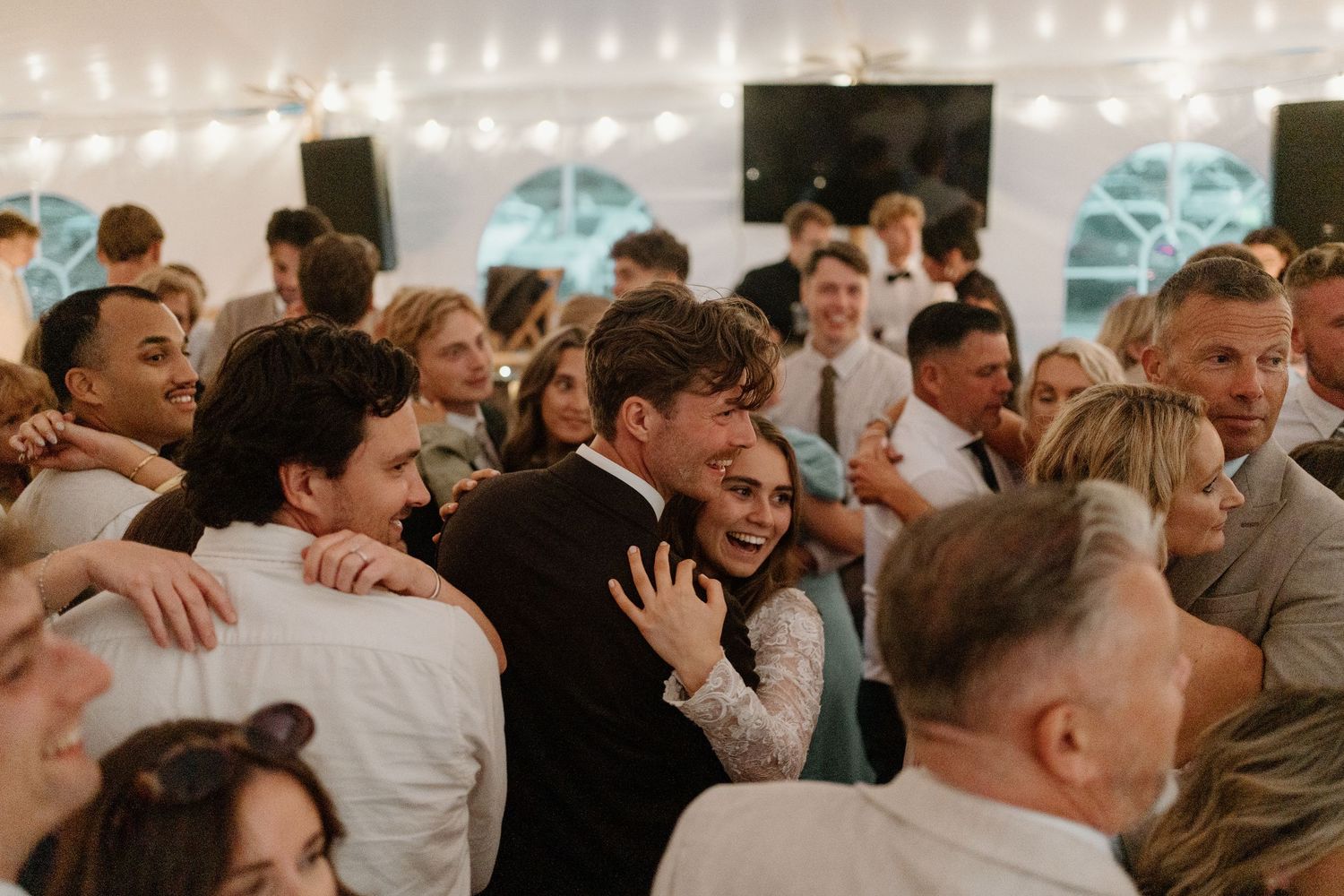 Wedding guests joyfully embracing couple during lively indoor reception celebration.