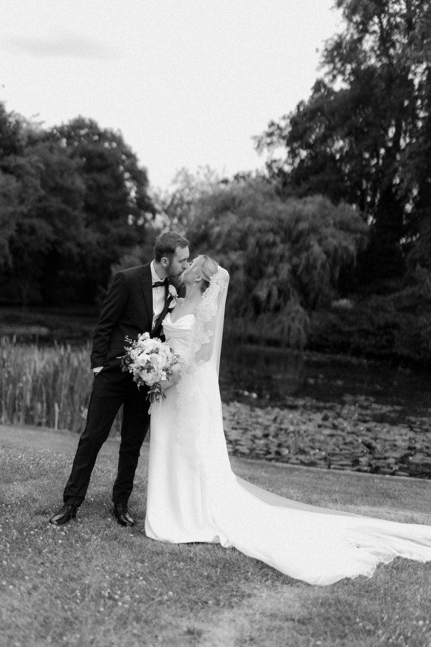 Black and white romantic photo of bride and groom kissing beside tranquil lake with willow trees.