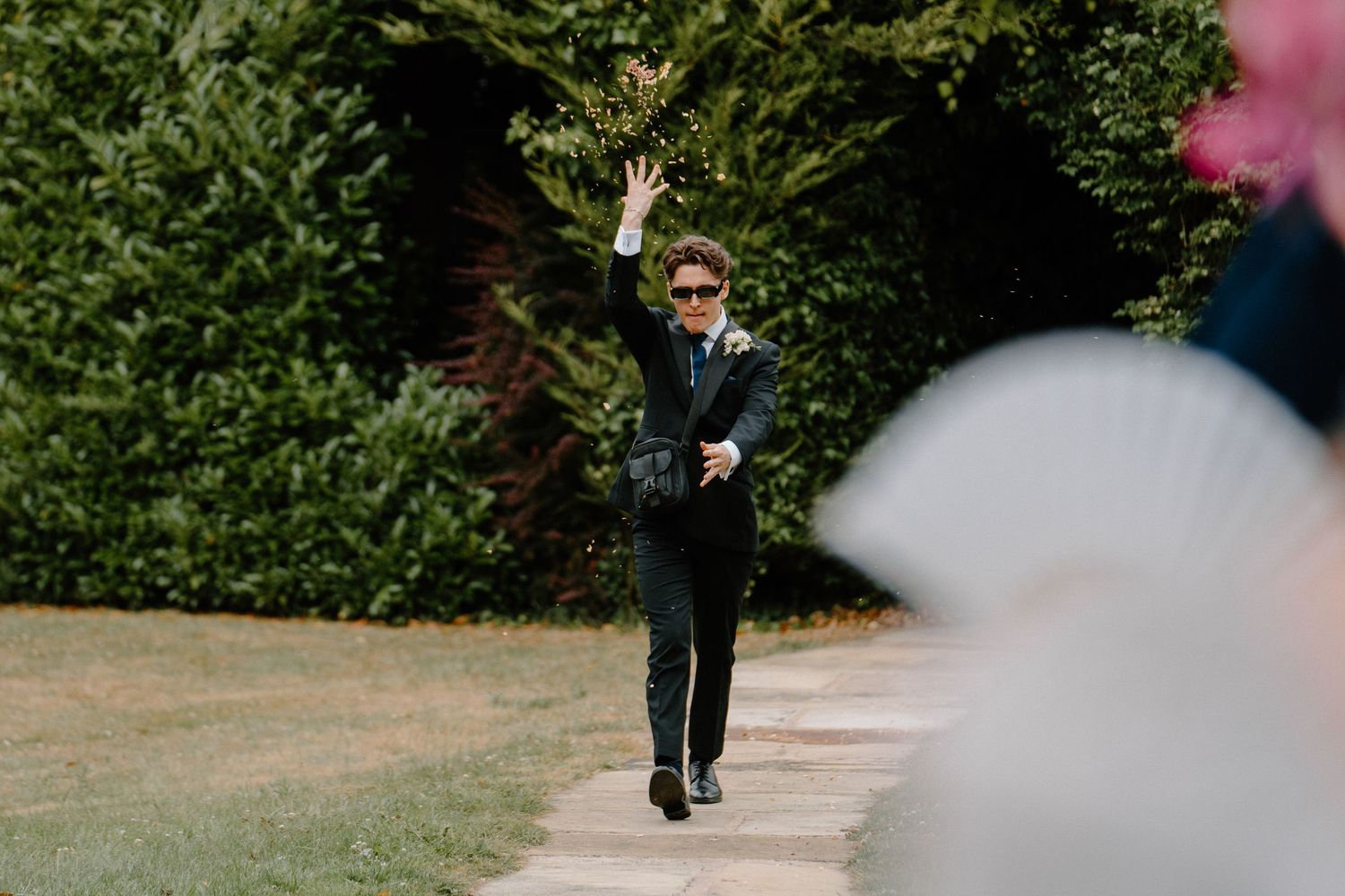Young man in dark suit and sunglasses waves while walking along garden pathway at outdoor wedding.