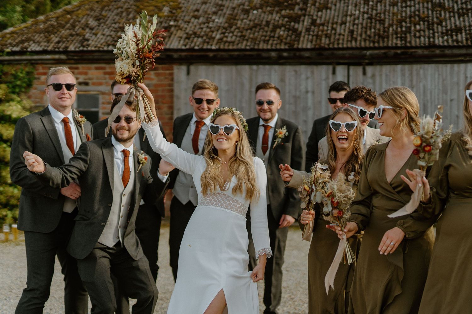 Joyful bride in sleek white gown raises bouquet while wedding party in olive attire and sunglasses celebrates.
