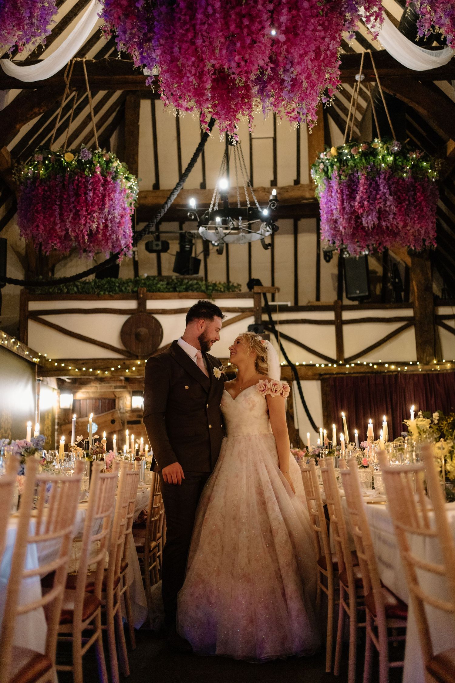 A bride and groom share a romantic moment beneath stunning purple floral installations in a barn venue.