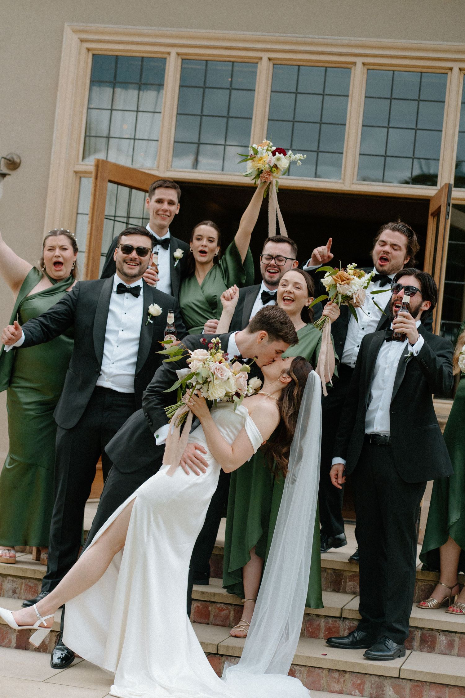 Bride and groom share a dip kiss surrounded by cheering wedding party on venue steps.