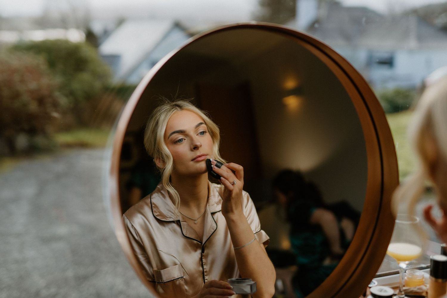 Bride applying lipstick reflected in round copper mirror during wedding morning preparations indoors.