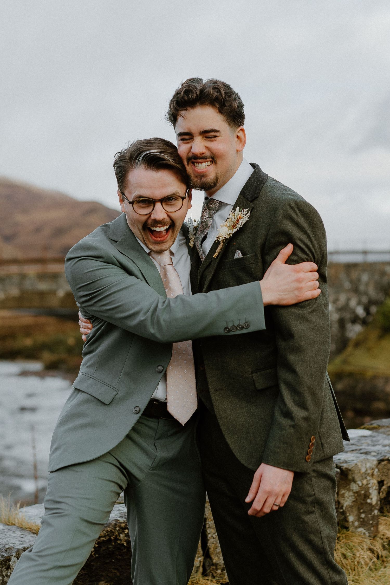 Two groomsmen in green suits laugh and embrace playfully on a Scottish riverside bridge.