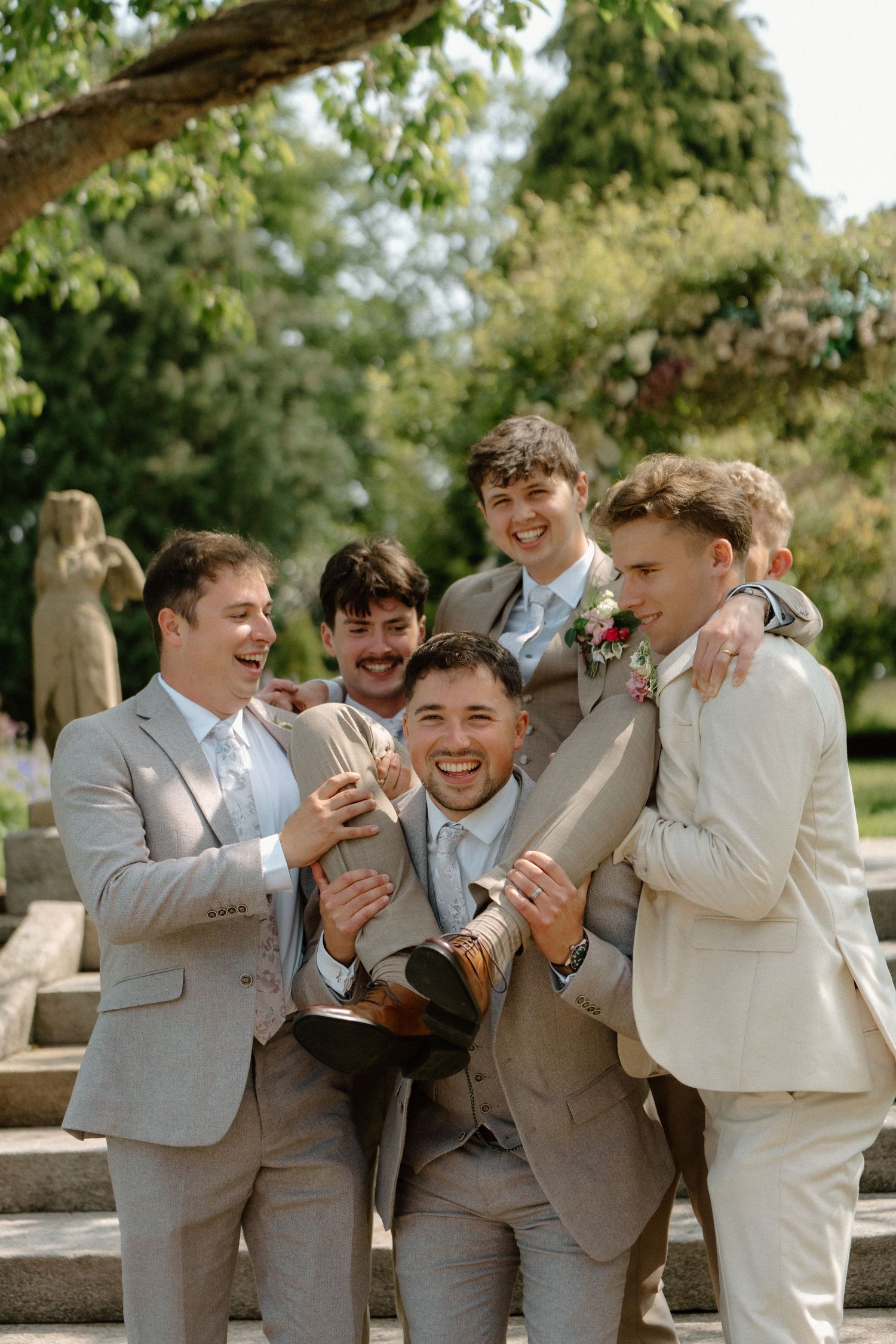 Laughing groom in grey suit lifted by smiling groomsmen in matching suits at outdoor wedding venue.