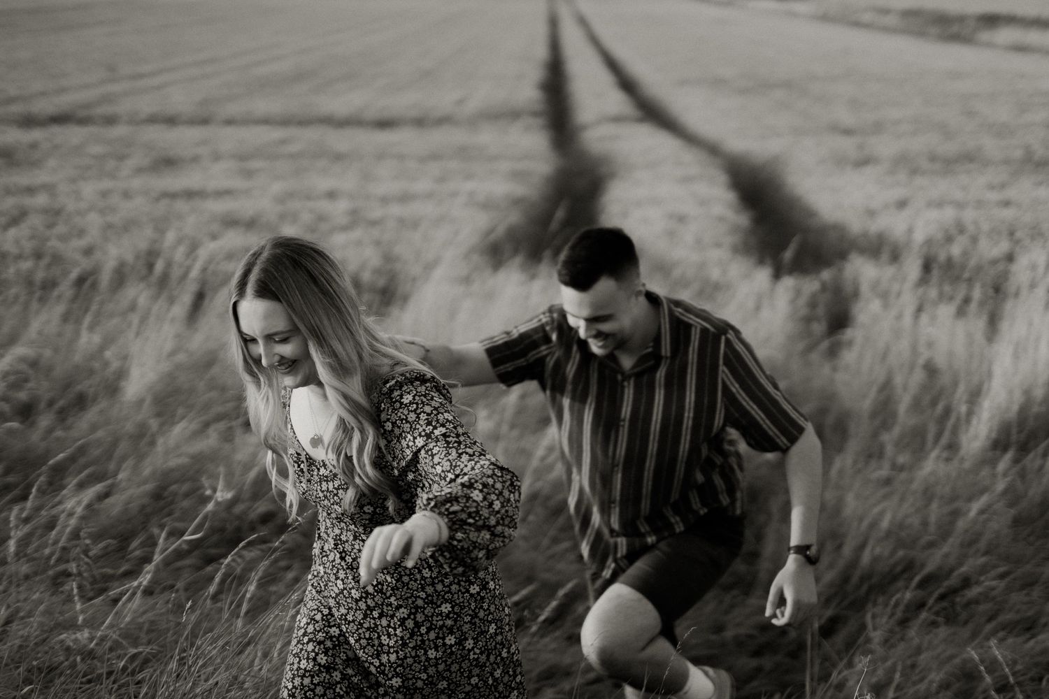 A couple runs playfully through a wind-swept field in black and white photography.