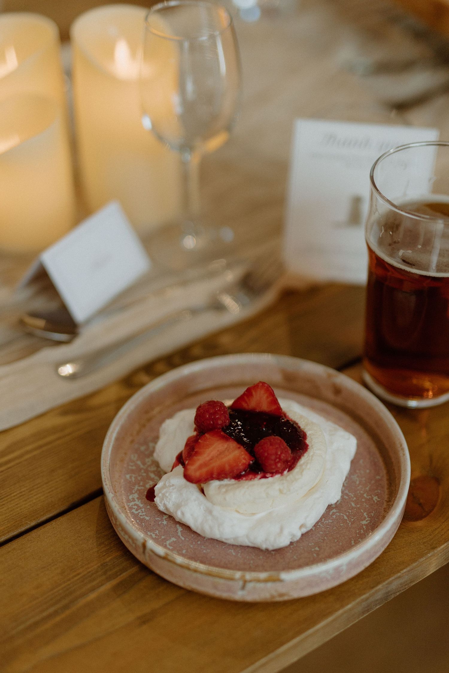 A pavlova dessert topped with fresh strawberries and berries served alongside a glass of dark beer on a wooden table.