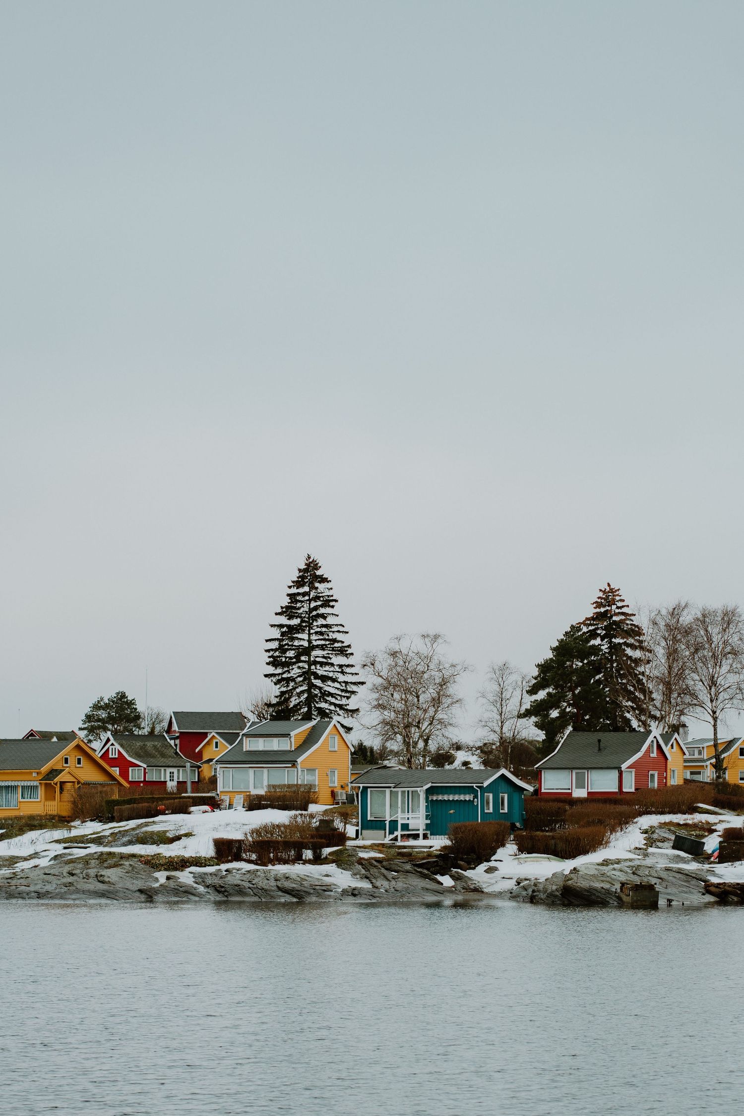 Colorful coastal houses line a snowy winter shoreline in a small fishing village with evergreen trees against a grey sky.
