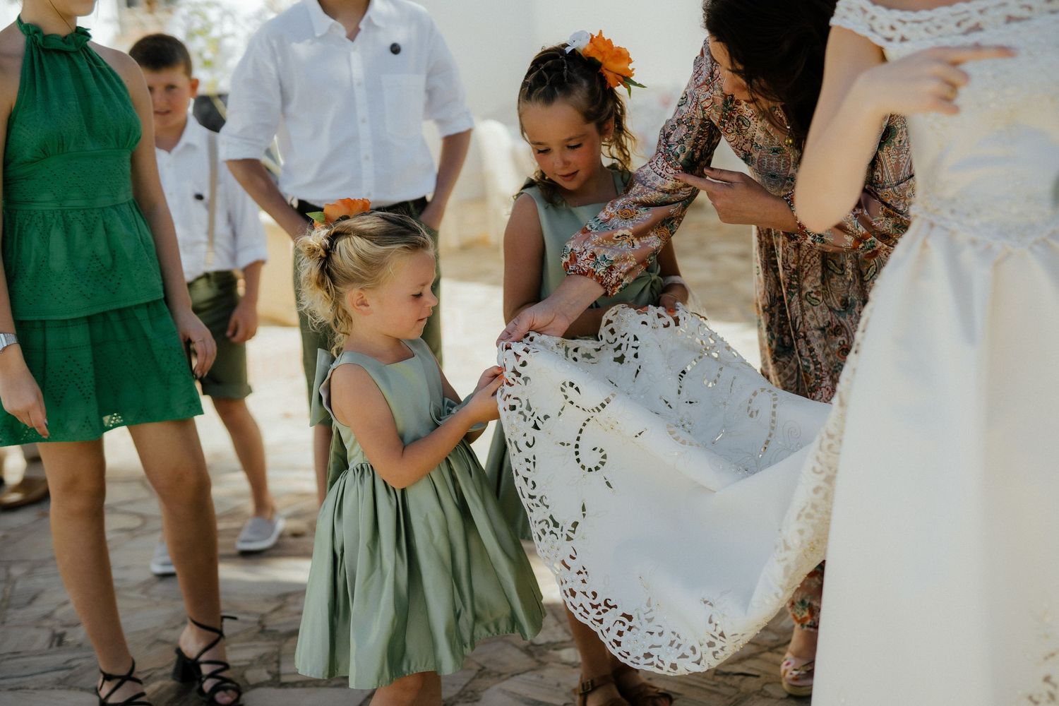 Two young flower girls in sage green dresses help arrange the lace hem of a bride's wedding gown outdoors.