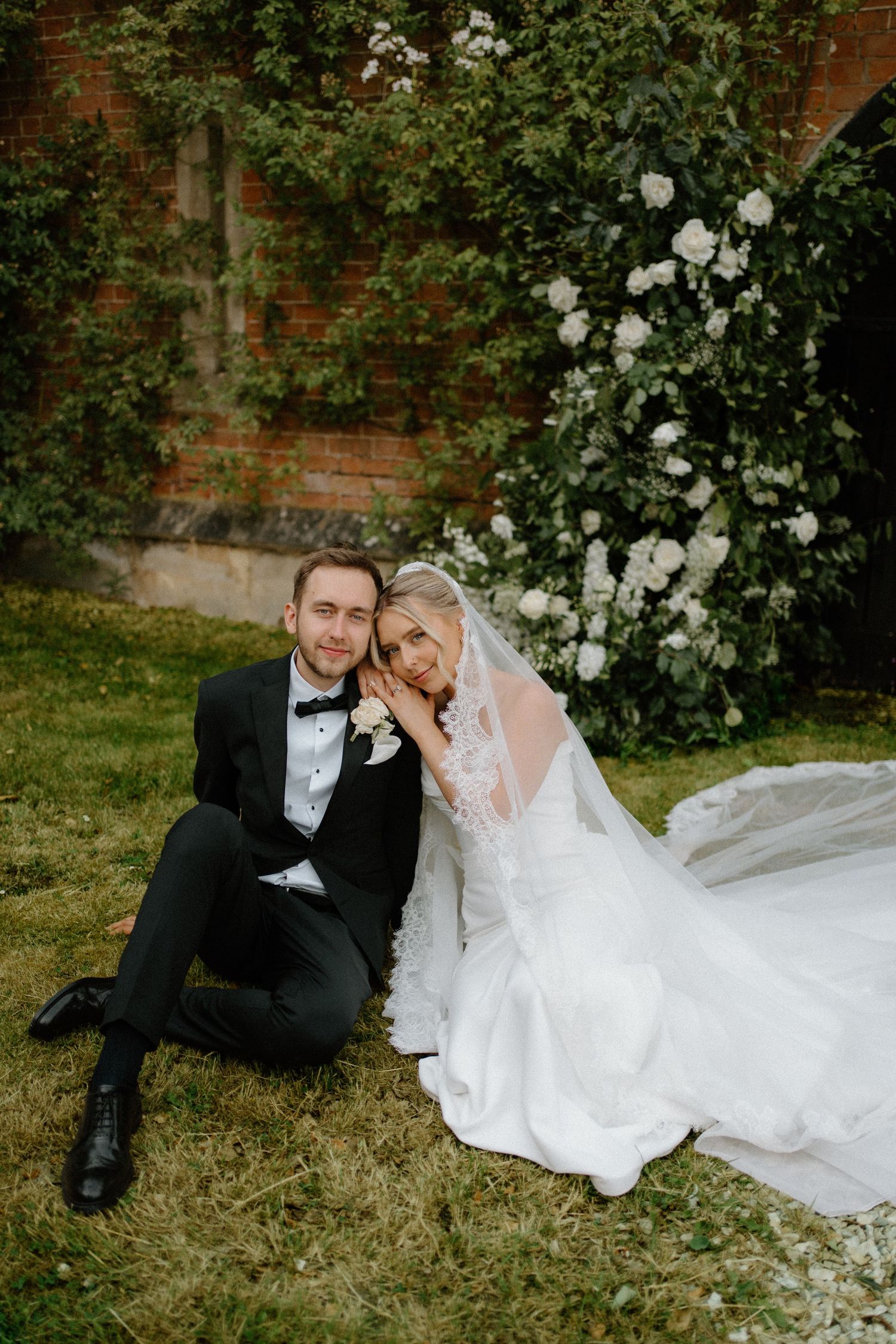 Bride and groom sitting on grass beside white roses and brick wall in a romantic outdoor wedding portrait.