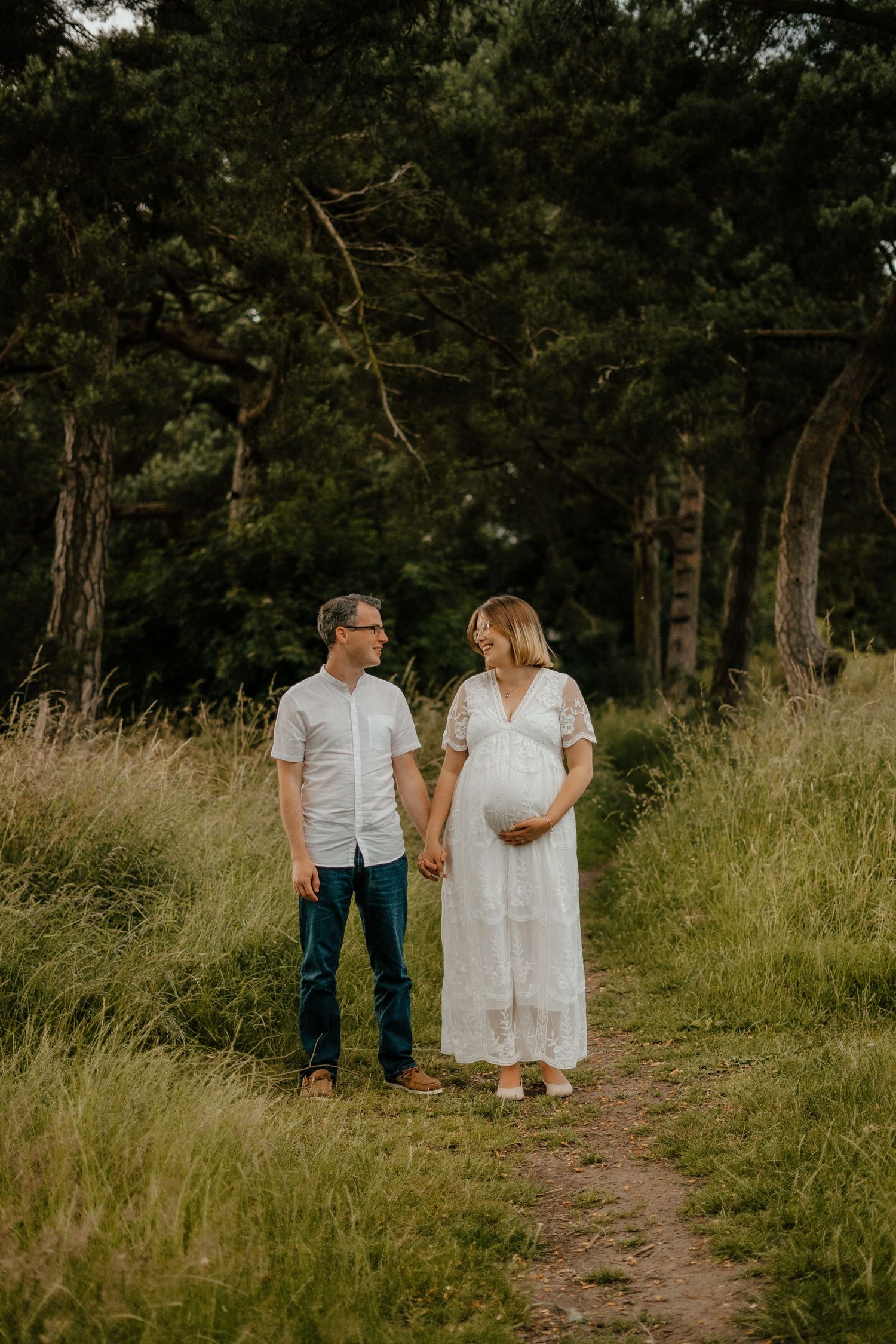 A pregnant couple holds hands on a woodland path, smiling at each other lovingly.