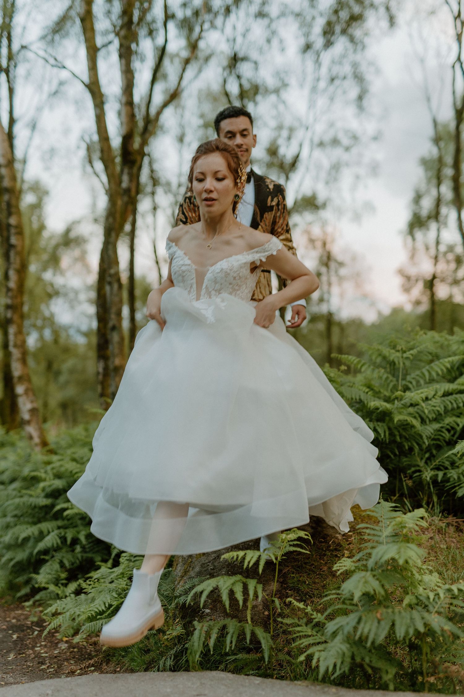 Bride in off-shoulder lace wedding gown walks through forest ferns with groom in gold patterned blazer behind her.