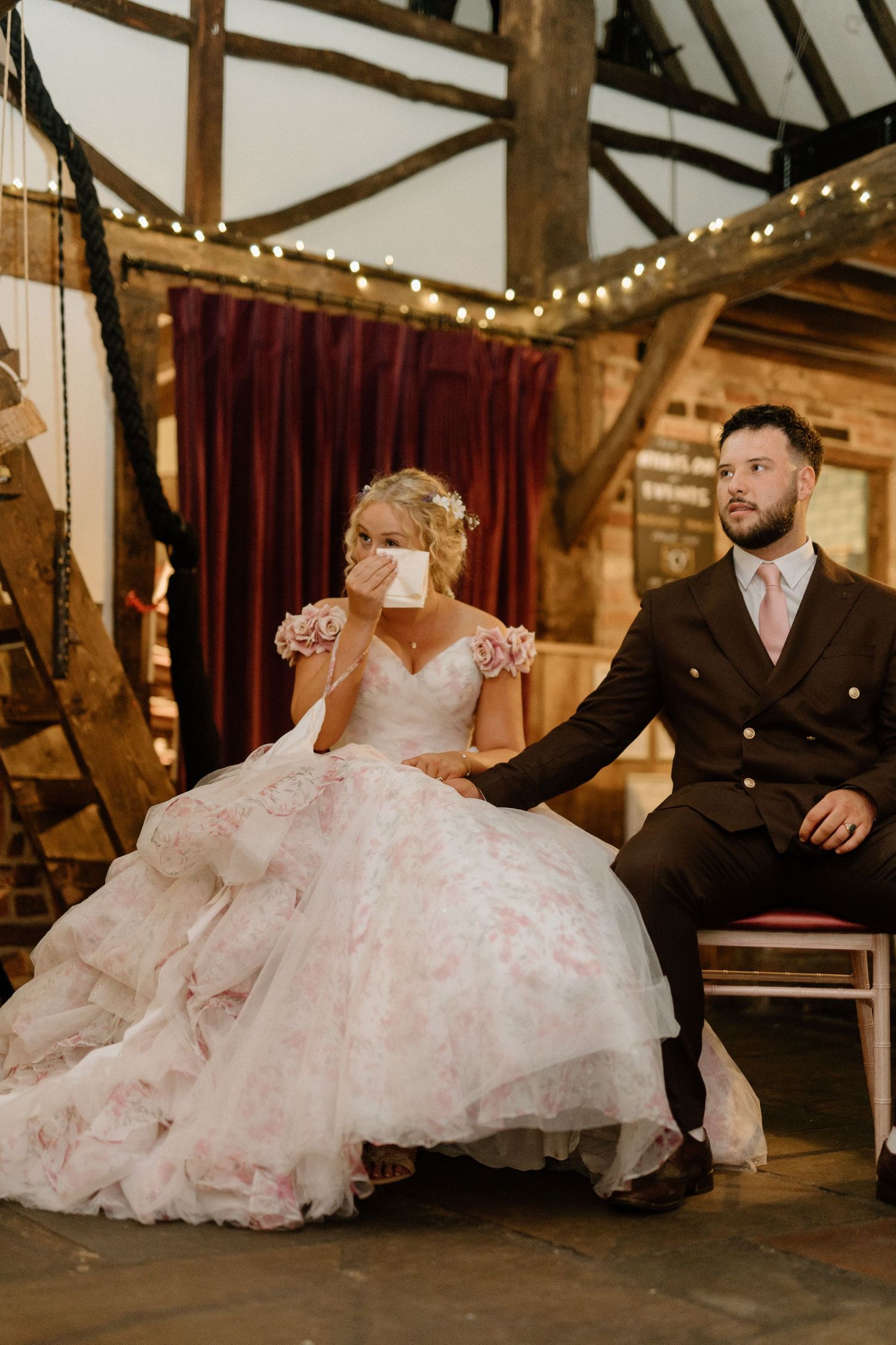 Bride in floral gown dabs tears while groom sits beside her during wedding speeches in a rustic barn venue.
