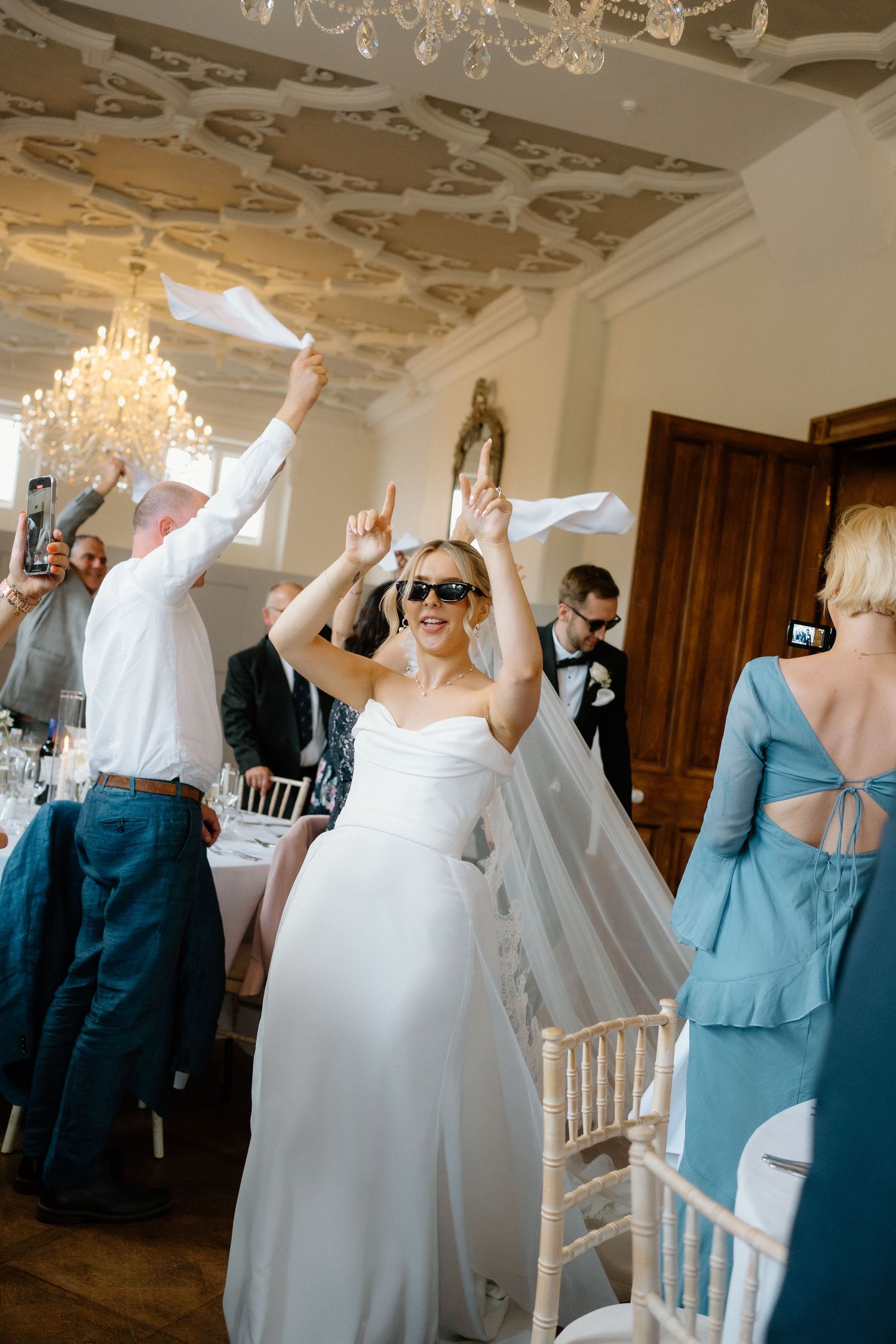 Bride in white gown wearing sunglasses making a grand entrance into the wedding reception with guests cheering.