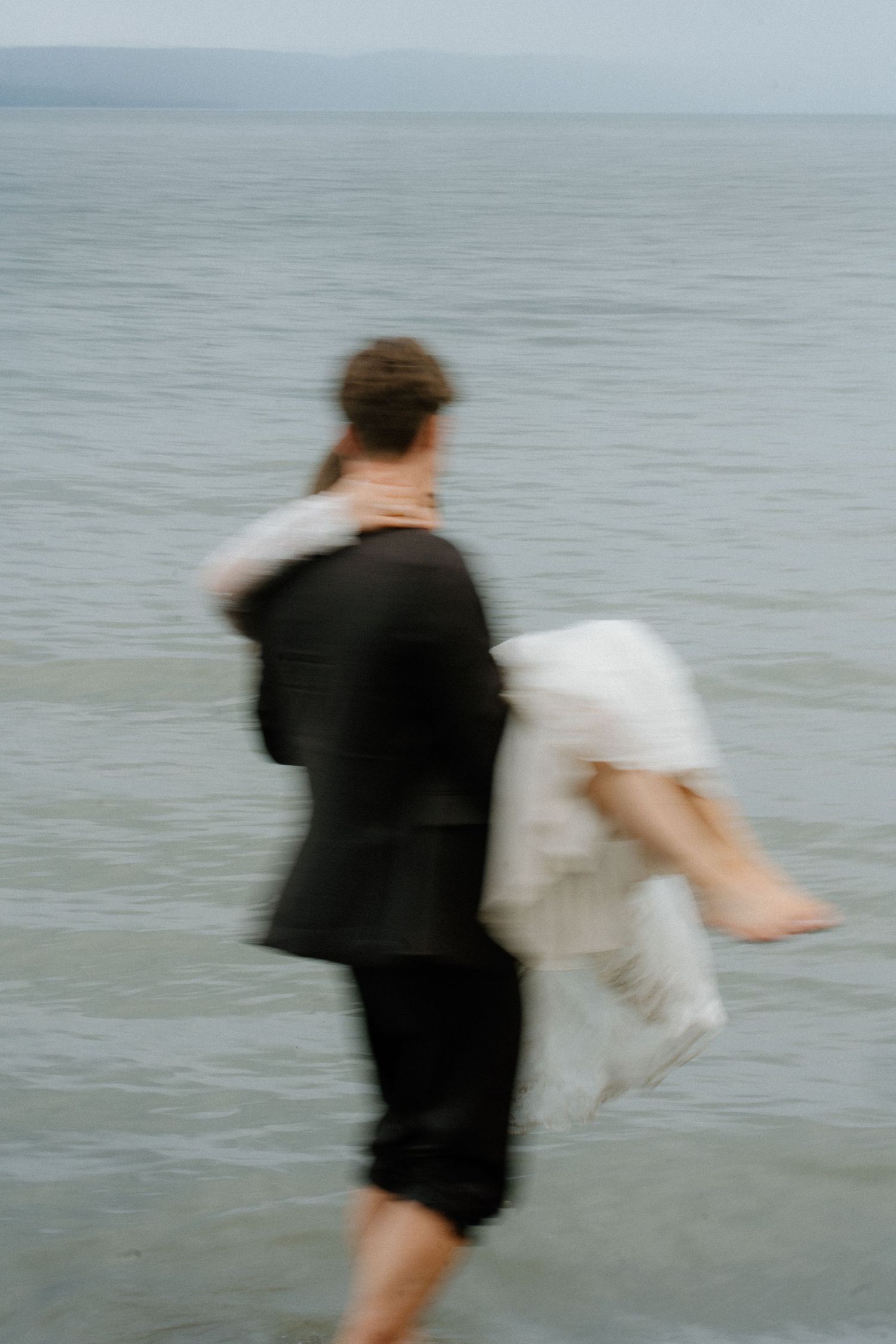 Motion blur artistic shot of groom carrying barefoot bride into ocean waves.