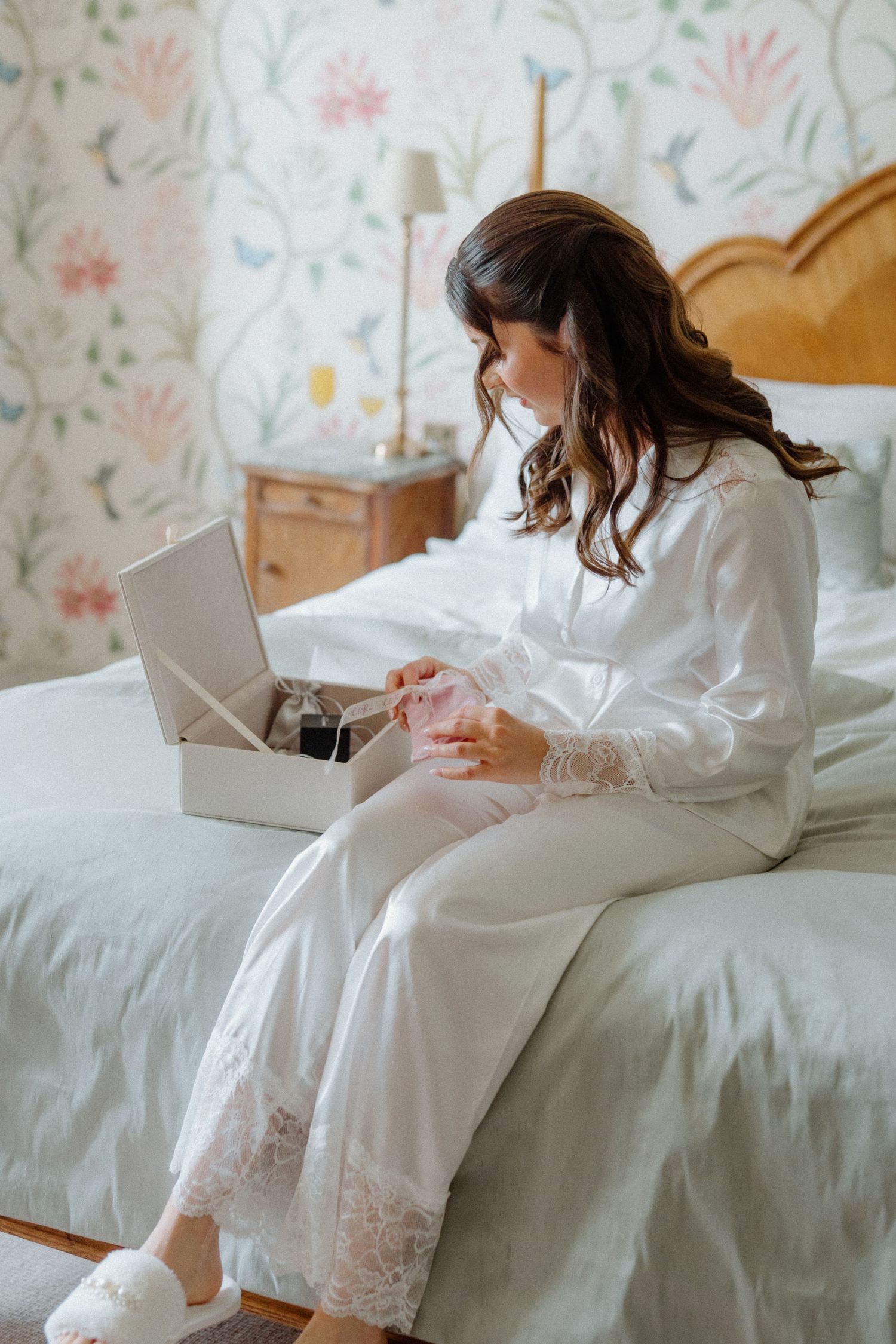 Bride in white silk robe sitting on bed opening a gift box on wedding morning.