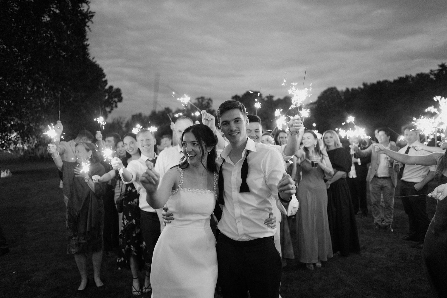 Black and white photo of bride and groom walking through sparkler exit with wedding guests at dusk.