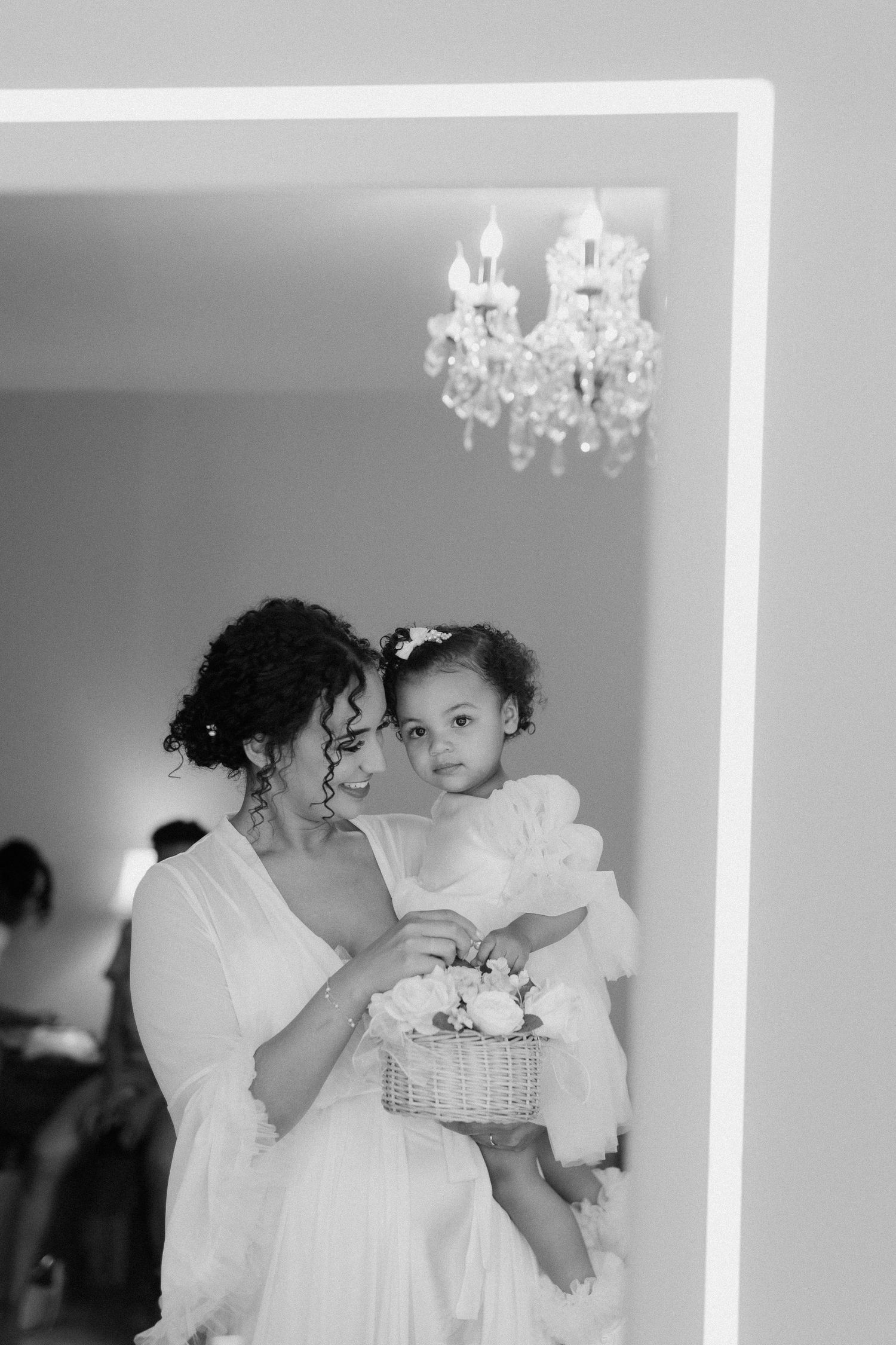 Bride holding a young flower girl with a basket of flowers, reflected in a mirror.