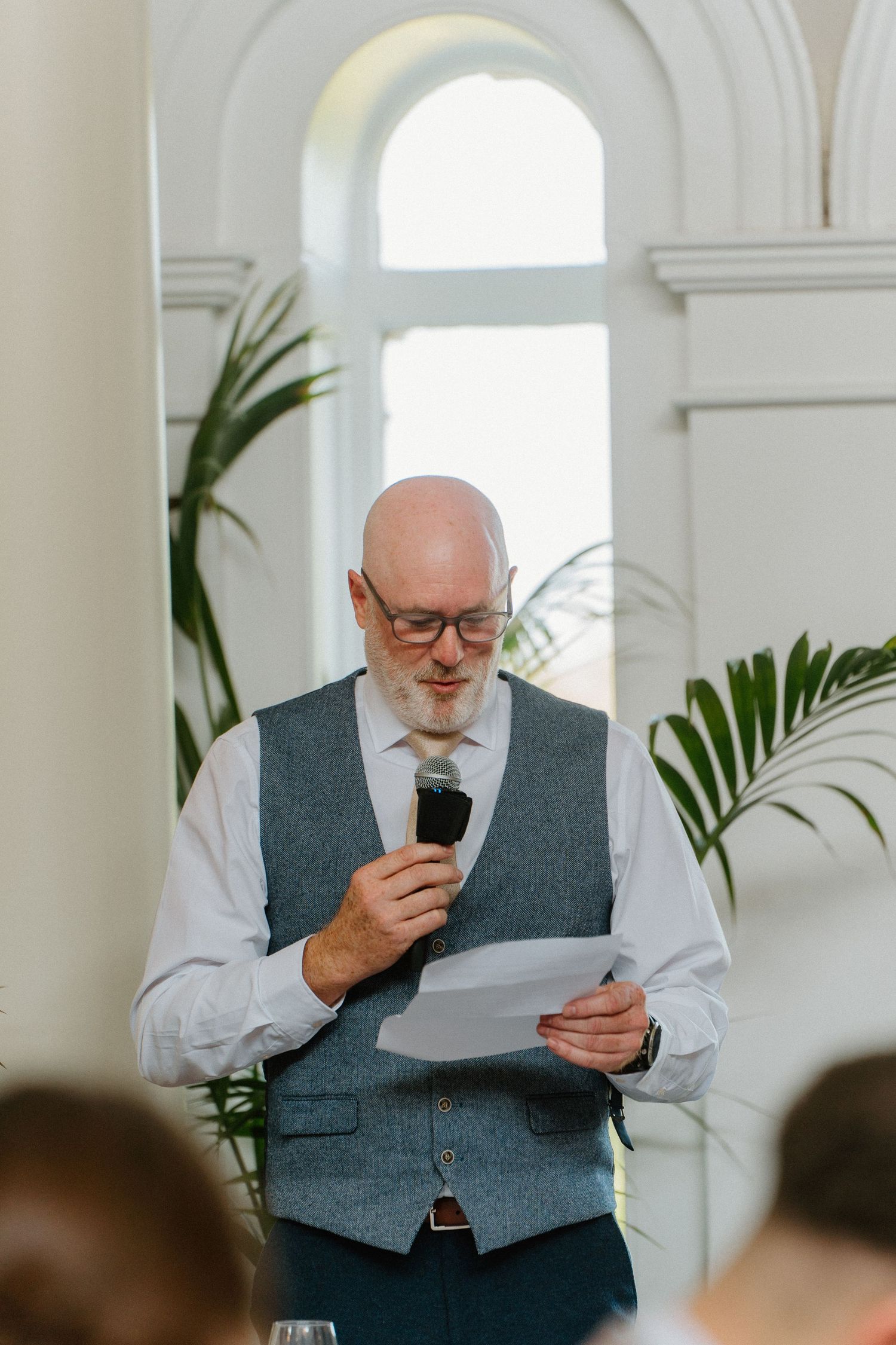 Bald man with glasses delivering a wedding speech while holding a microphone and paper in an elegant white venue.