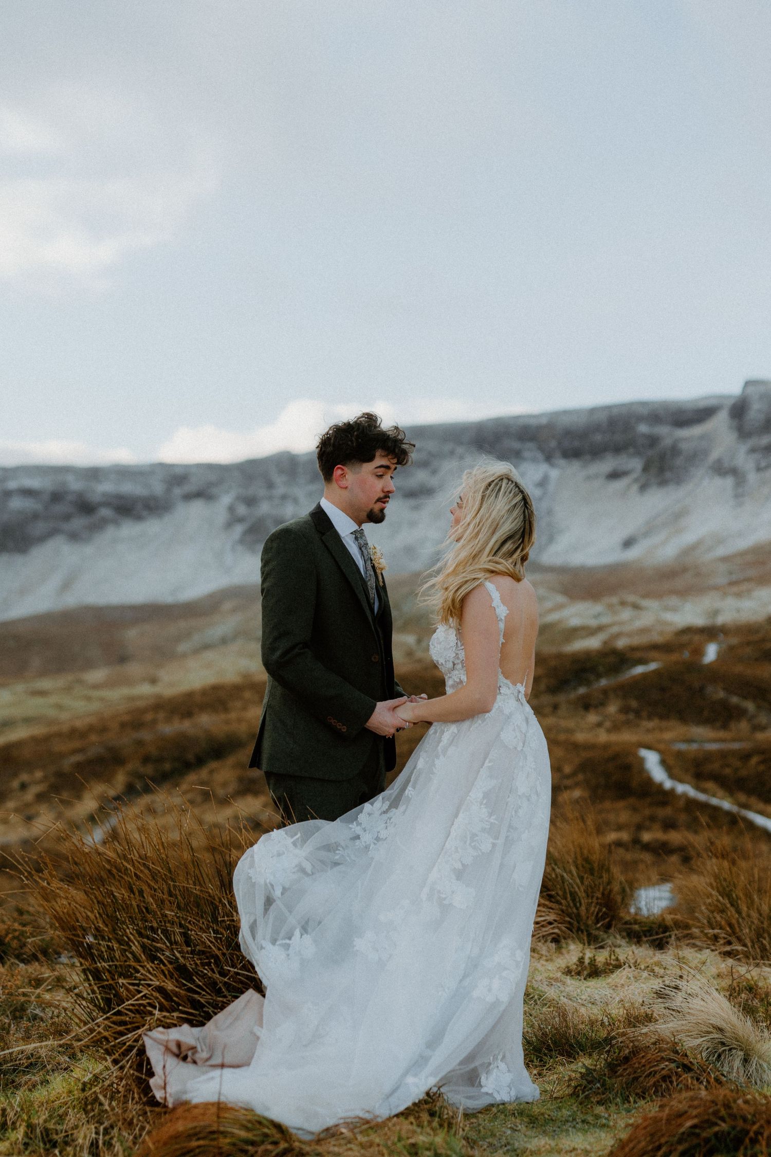 Bride in flowing lace gown holds hands with groom in green tweed suit in Scottish Highlands.